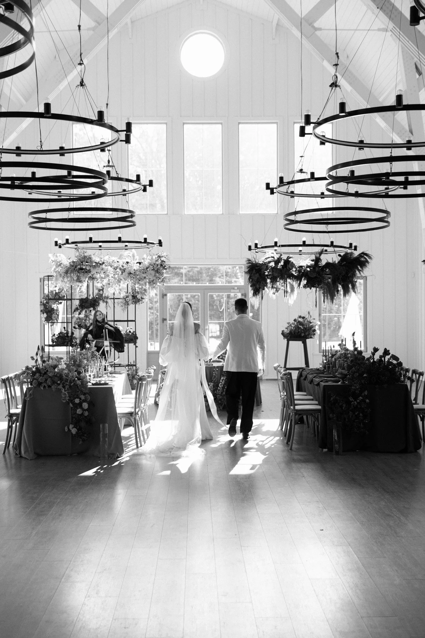 A bride and groom walking hand in hand inside a decorated banquet hall with large windows, floral arrangements, and chandeliers.
