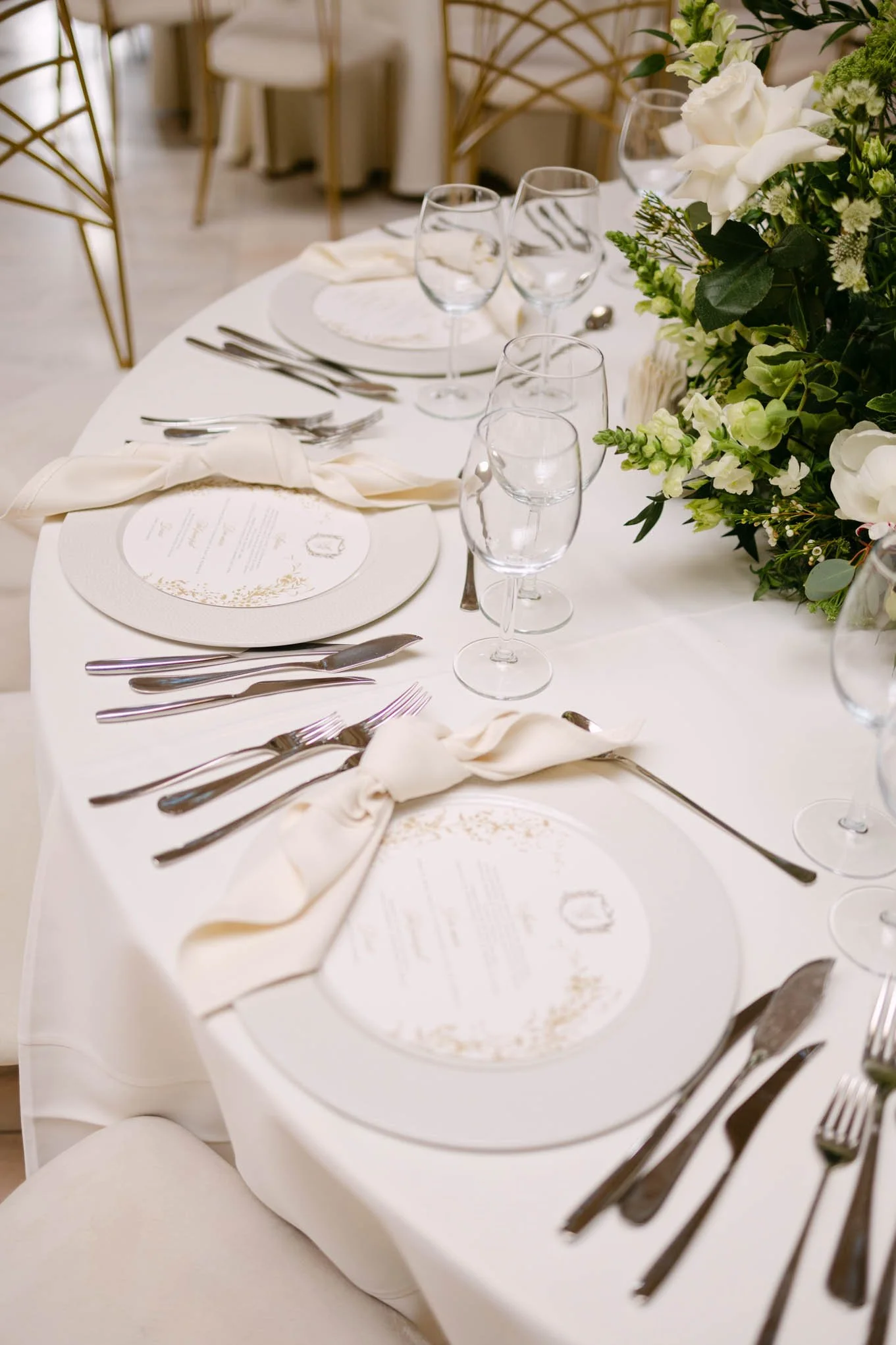 Elegant wedding reception table setting with white tablecloth, floral centerpiece, white napkins tied with ribbons, clear wine glasses, and assorted silverware.