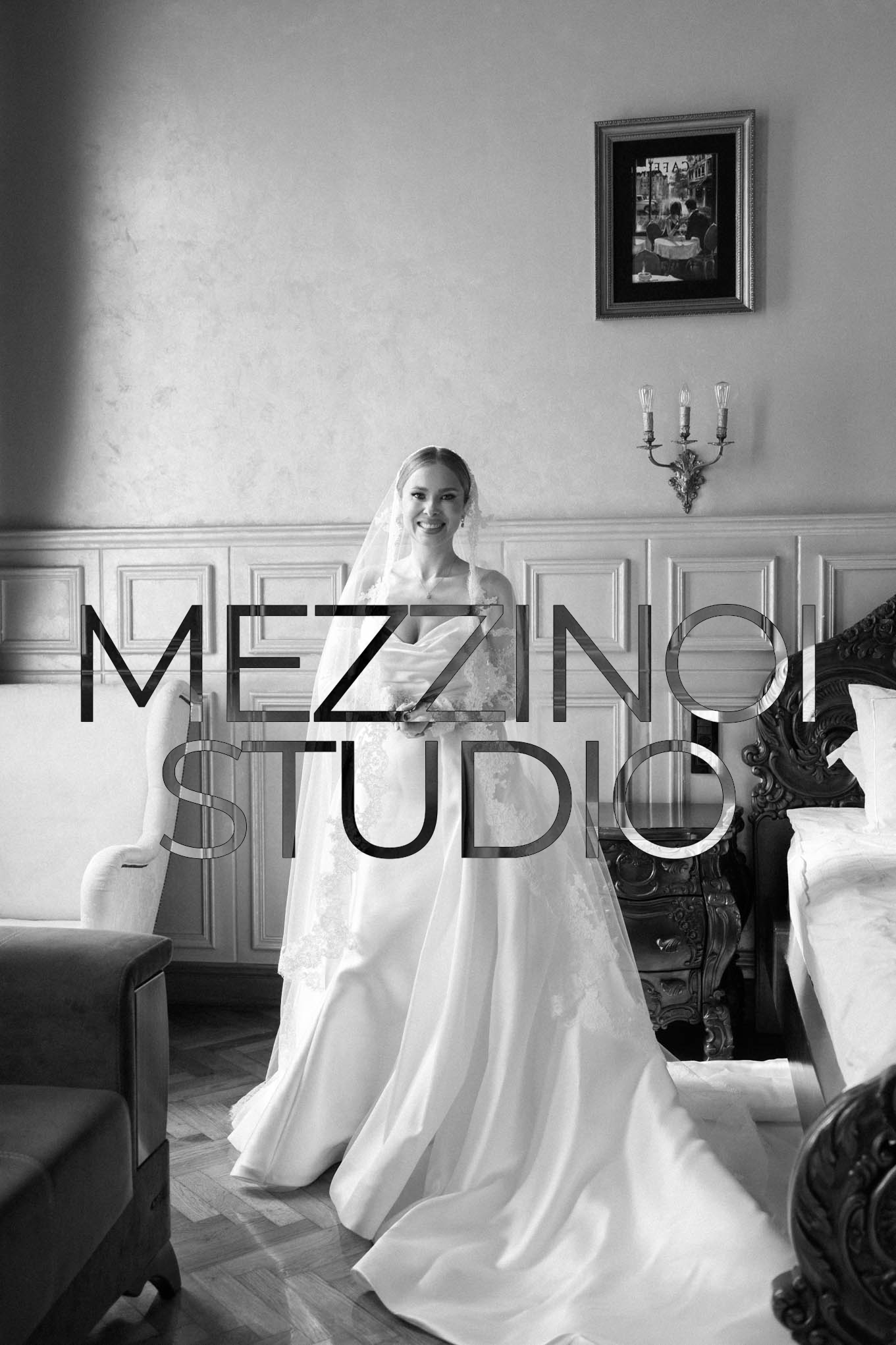 Black and white photo of a smiling bride in a wedding dress and veil inside a room with ornate furniture and framed artwork on the wall.