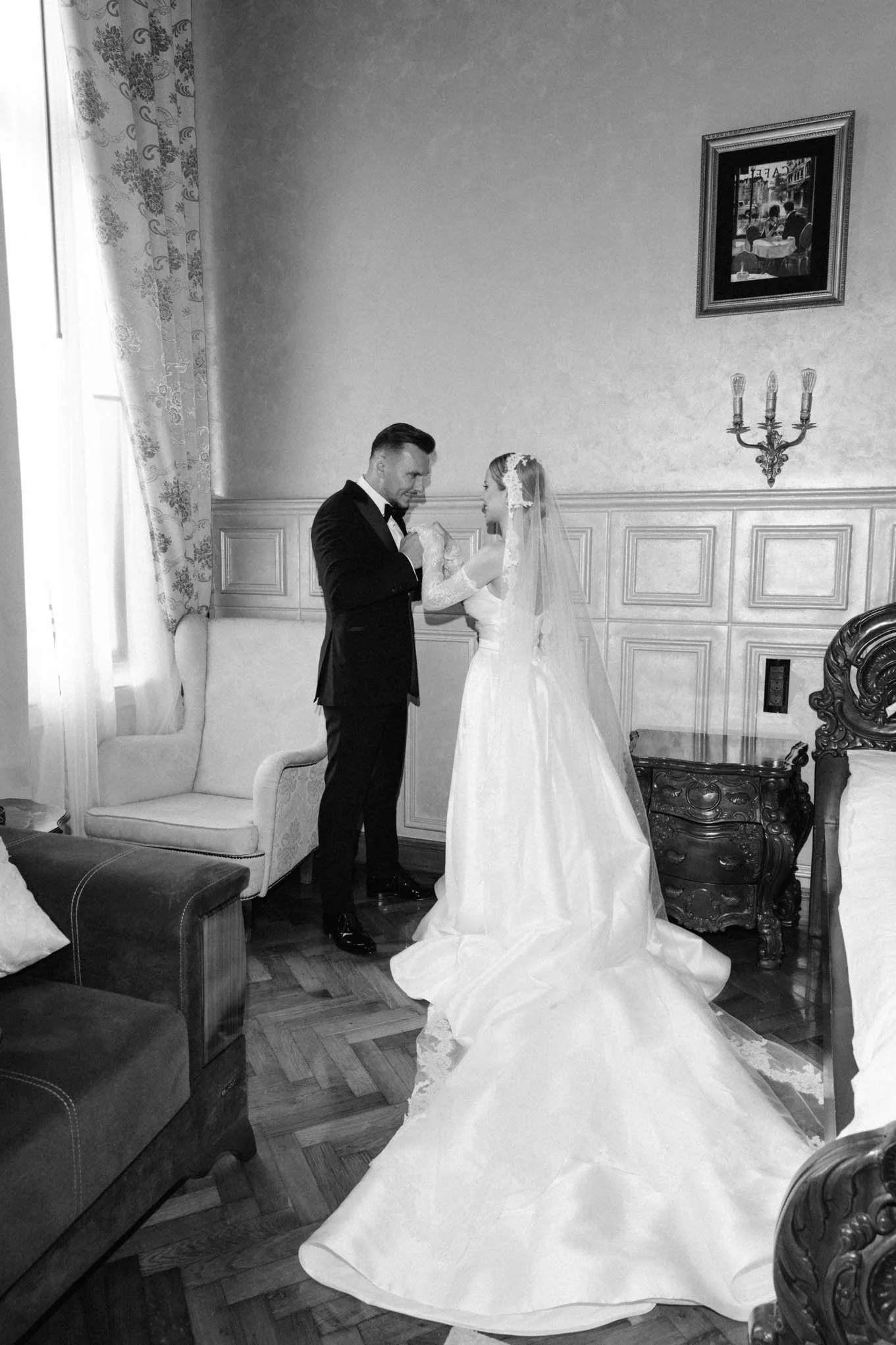 A black and white photo of a bride and groom standing inside a room. The groom, dressed in a tuxedo, and the bride, wearing a wedding gown and veil, are facing each other, holding hands, and smiling. Italian style wedding as seeon in Harpers Bazaar!