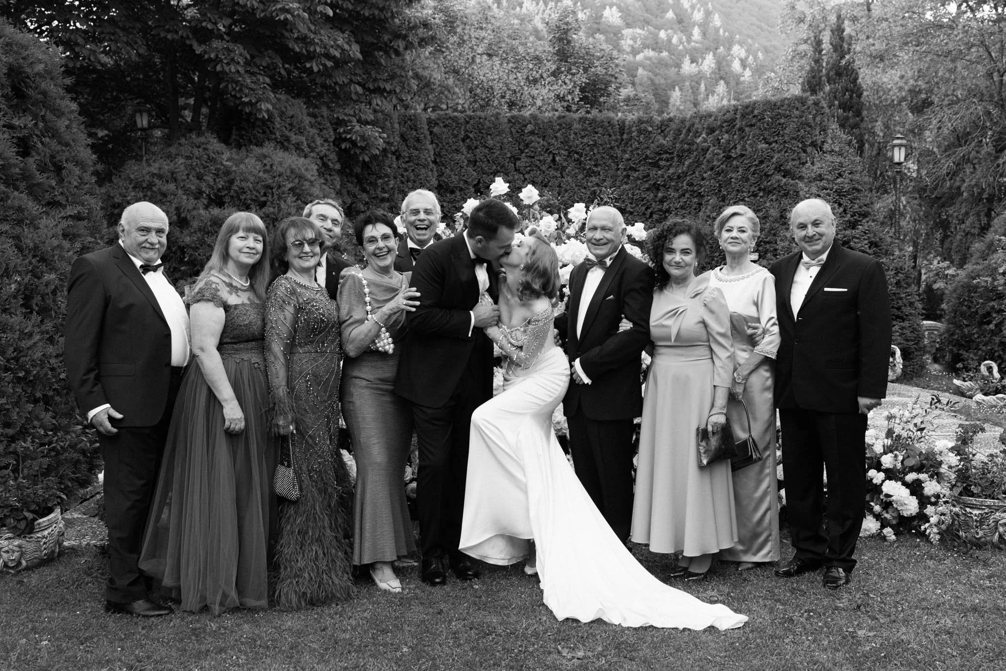 A wedding party in formal attire standing outdoors, with a bride and groom kissing in the center surrounded by family and friendsn on a Tuscany style garden Villa!