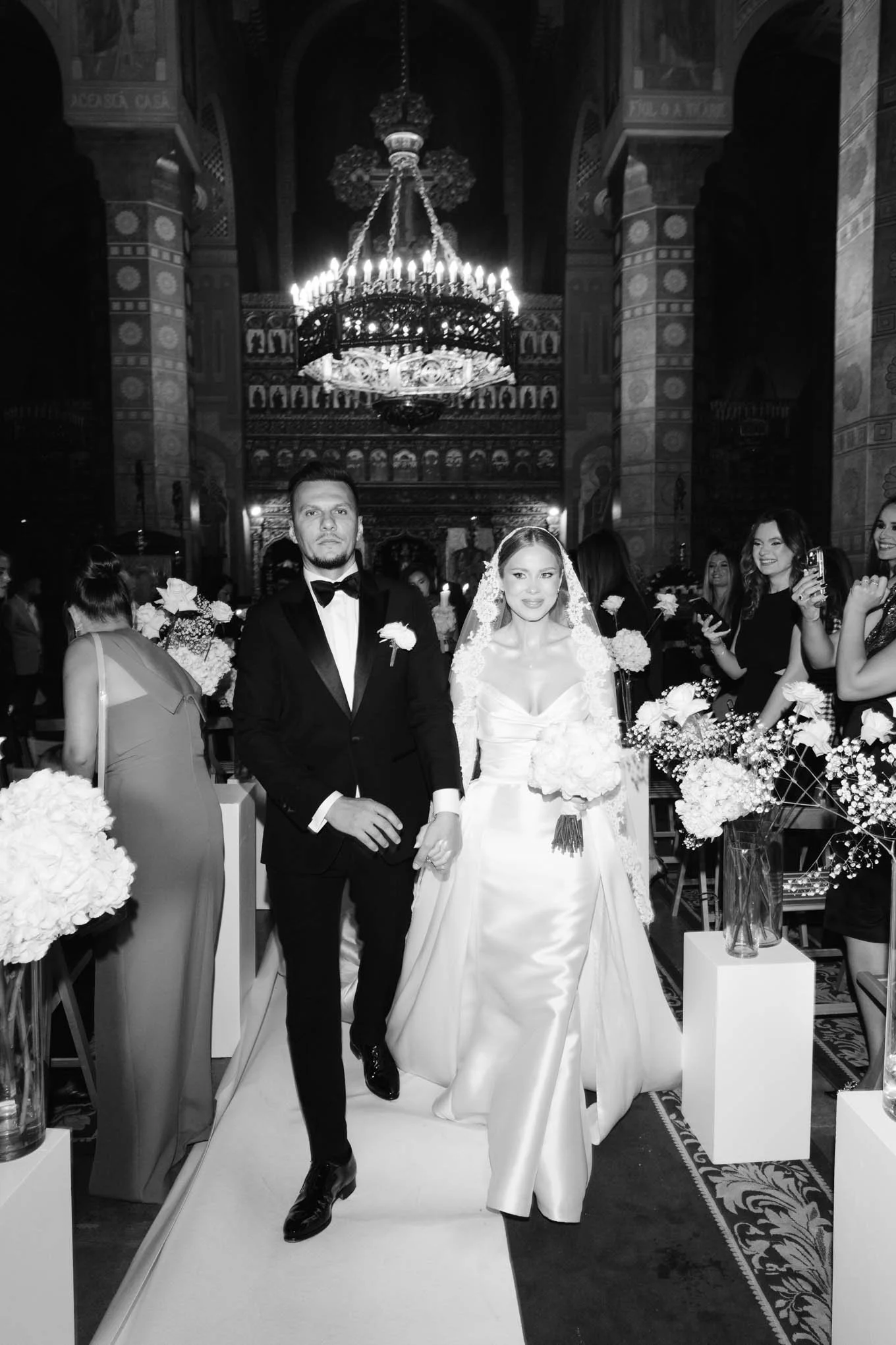 A bride and groom walking down the aisle at their wedding ceremony inside a decorated church with floral arrangements and chandeliers, surrounded by guests. As seen in vogue weddings, french weddings of south france!