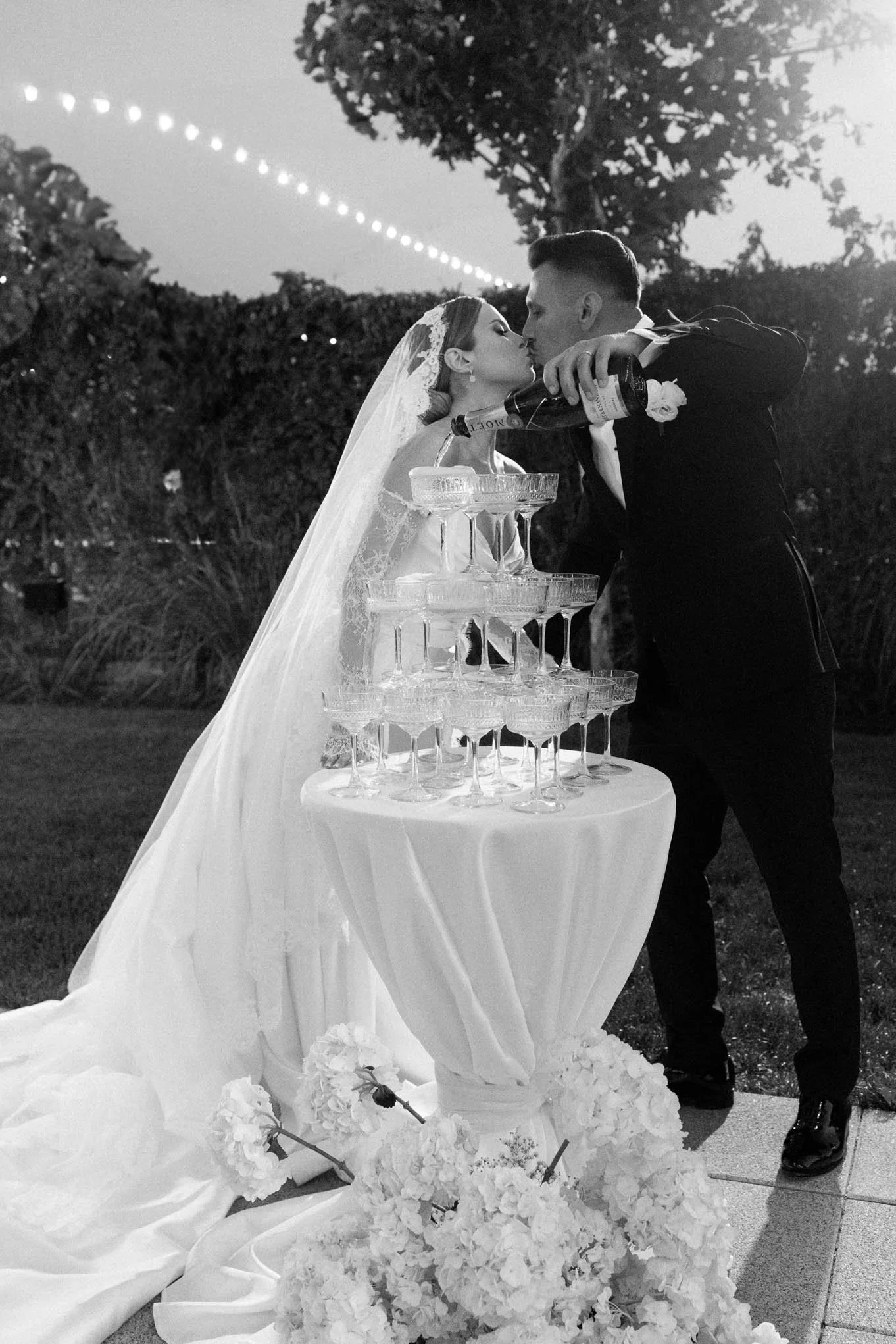 Bride and groom pouring champagne into a tower of glasses at their wedding reception outdoors at night. As seen in Vogue weddings, Bazaar weddings!