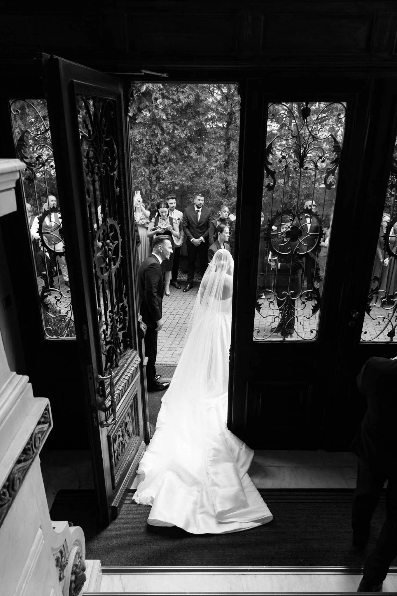 Black and white photo of a bride and groom standing at the open door of a church, with wedding guests outside. A frame as seen in vogue weddings!