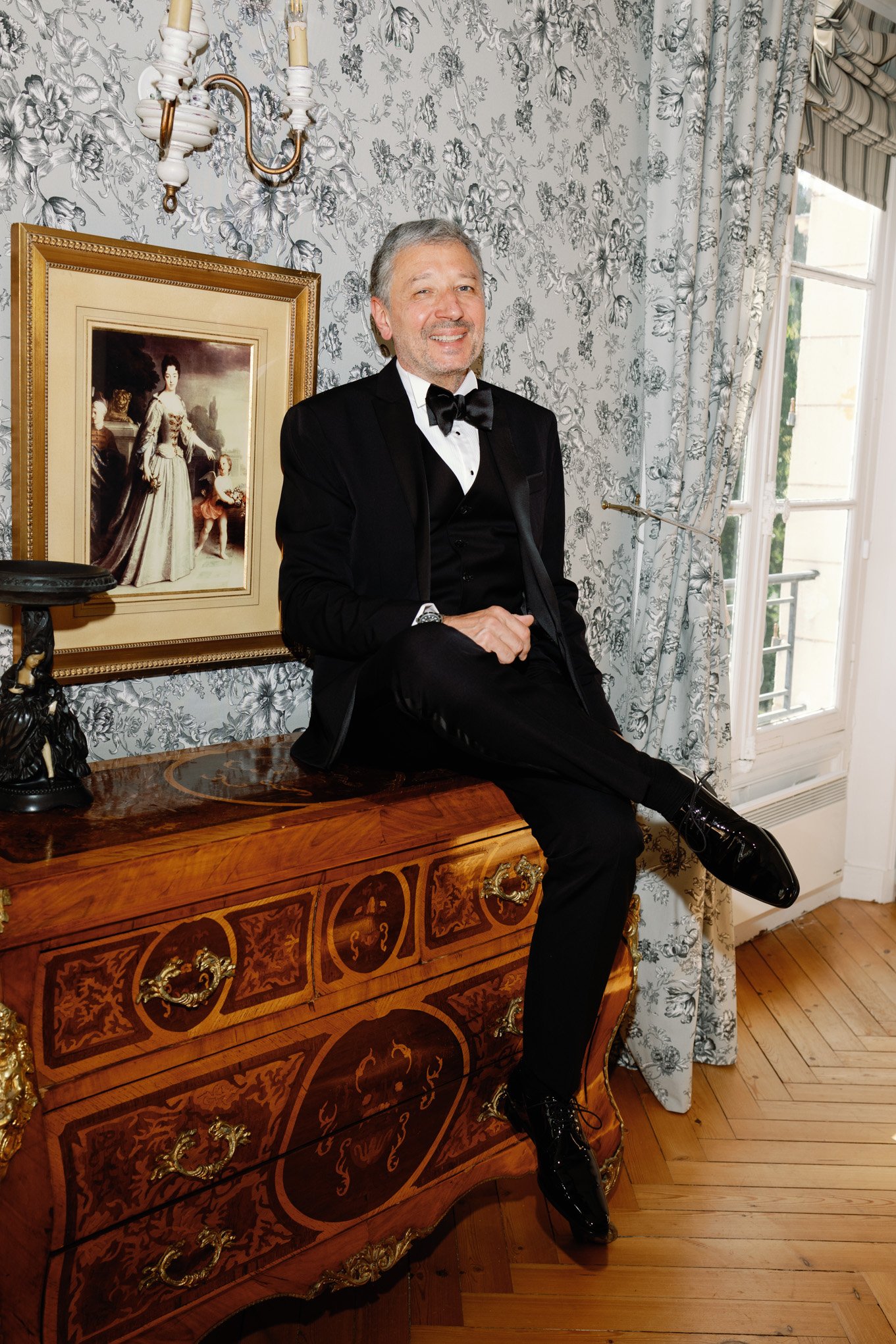 A man dressed in a tuxedo with a bow tie, sitting on a vintage wooden dresser, smiling, in an elegant room with floral wallpaper, a framed portrait of a woman and children, and a window with curtains.
