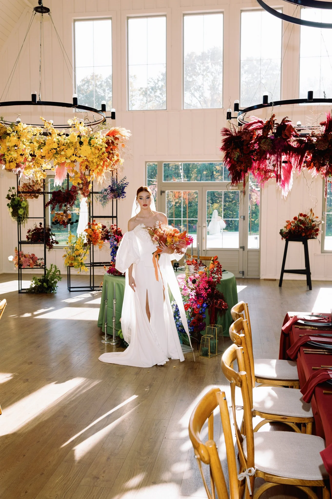 A woman in a white wedding dress holding a bouquet of flowers inside a decorated venue with floral arrangements hanging from black rings and a green tablecloth on a table filled with flowers, surrounded by wooden chairs with cream-colored cushions.