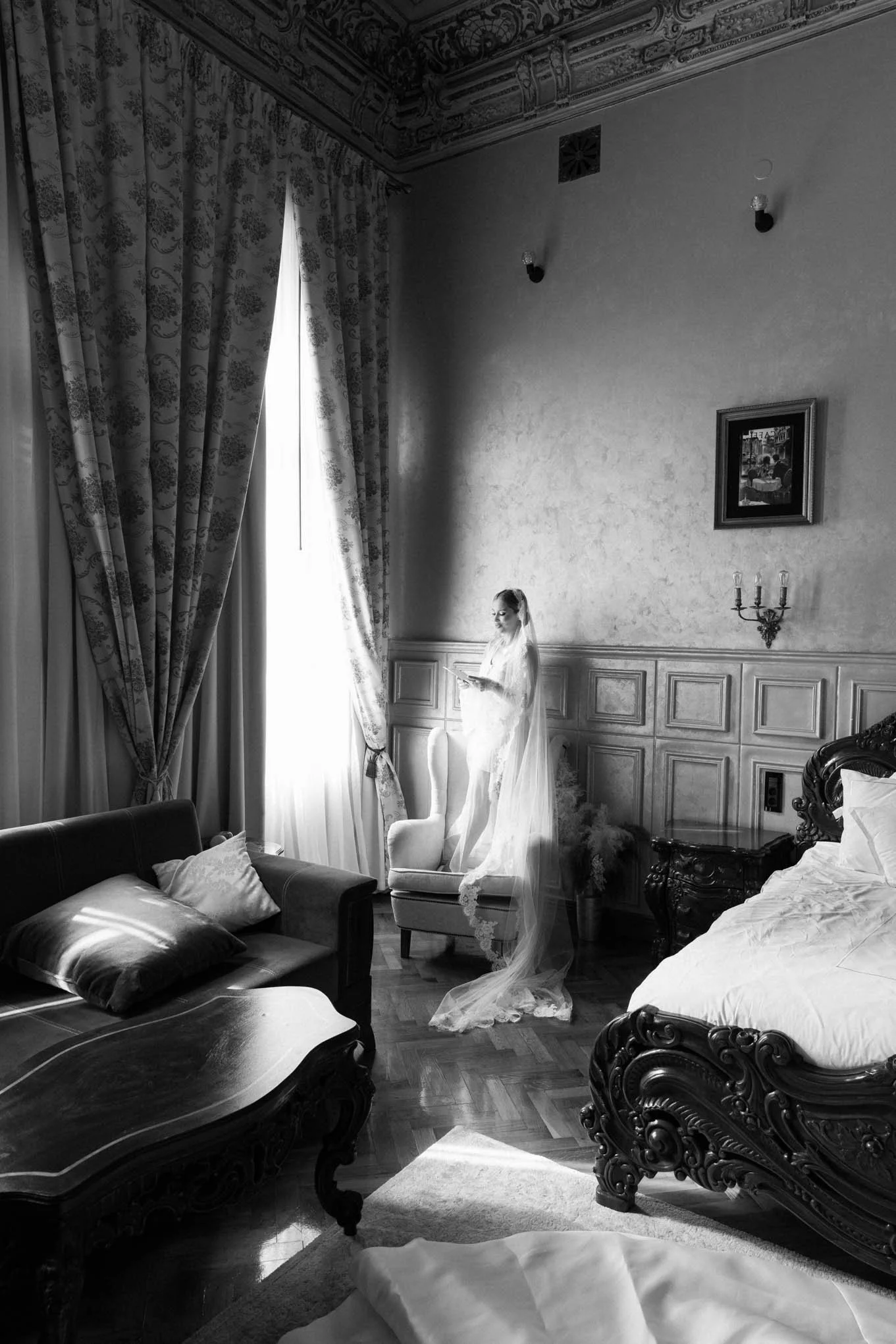 Black and white photo of a woman in a wedding dress and veil standing on a chair by a window, reading a book in a vintage, ornate bedroom with a large bed, a sofa, and curtains. Vogue style bride!