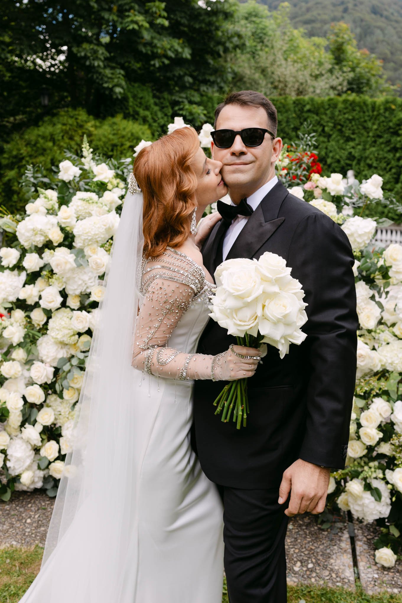 A bride with red hair in a wedding dress and veil kisses a groom in a black tuxedo, sunglasses, holding white roses, at a garden wedding, the style is italian aesthetic, tuscany style garden.