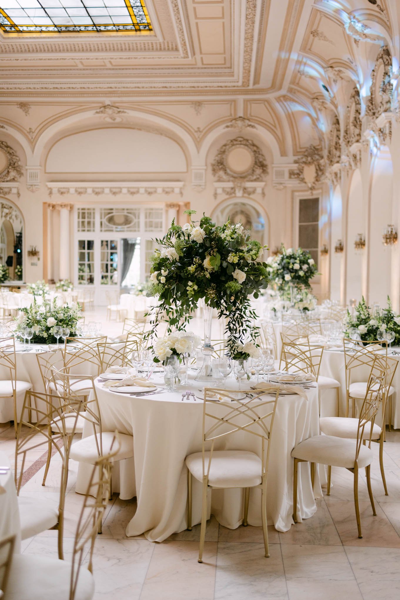Elegant banquet hall with round tables decorated with white tablecloths, tall floral centerpieces with white flowers and greenery, gold chairs, and soft lighting, suitable for a wedding reception or formal event.