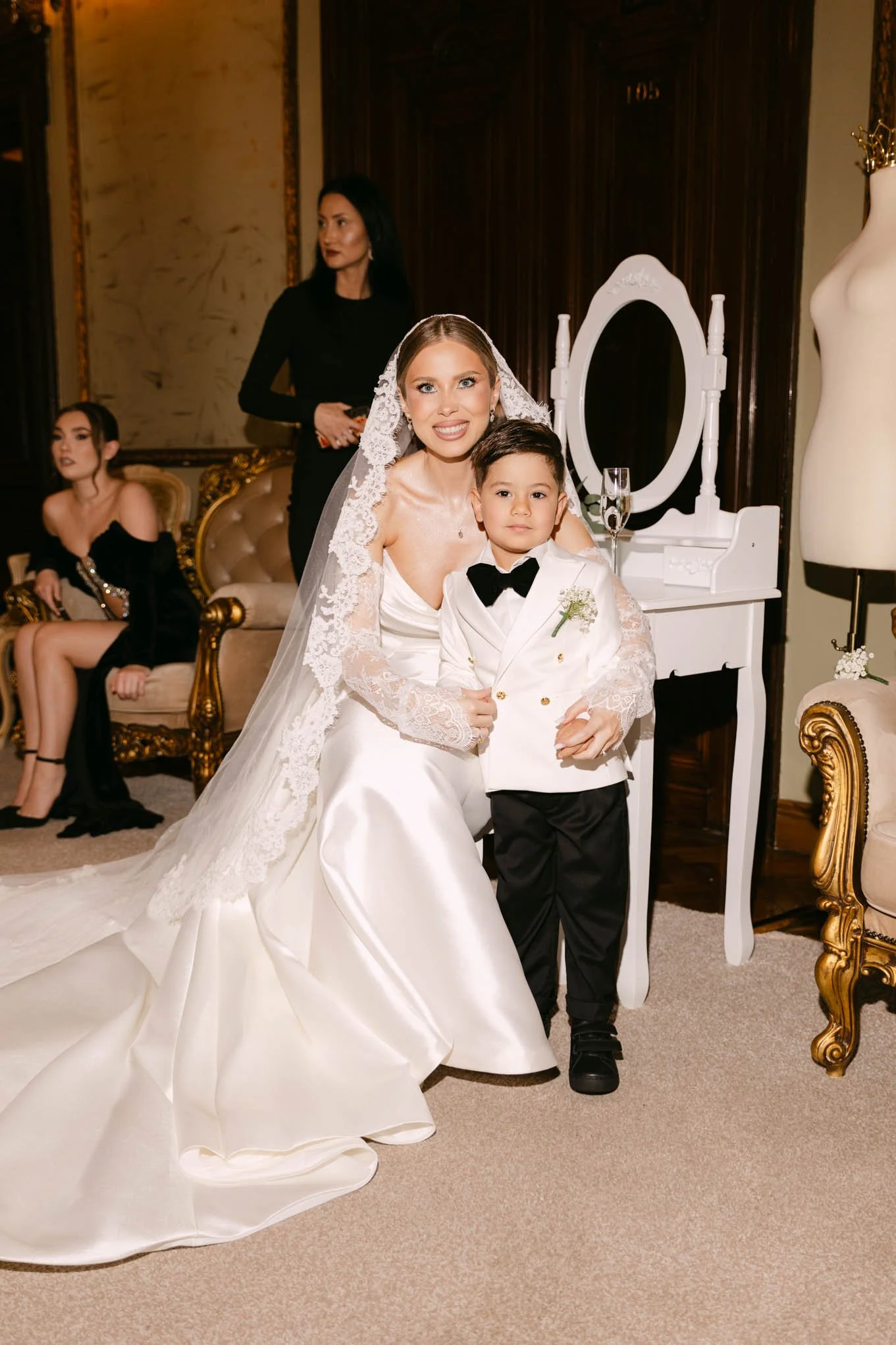 Bride in white wedding gown with lace veil smiling while sitting next to a young boy in a tuxedo, in a luxurious room with gold and white decor. Fashion vogue style!