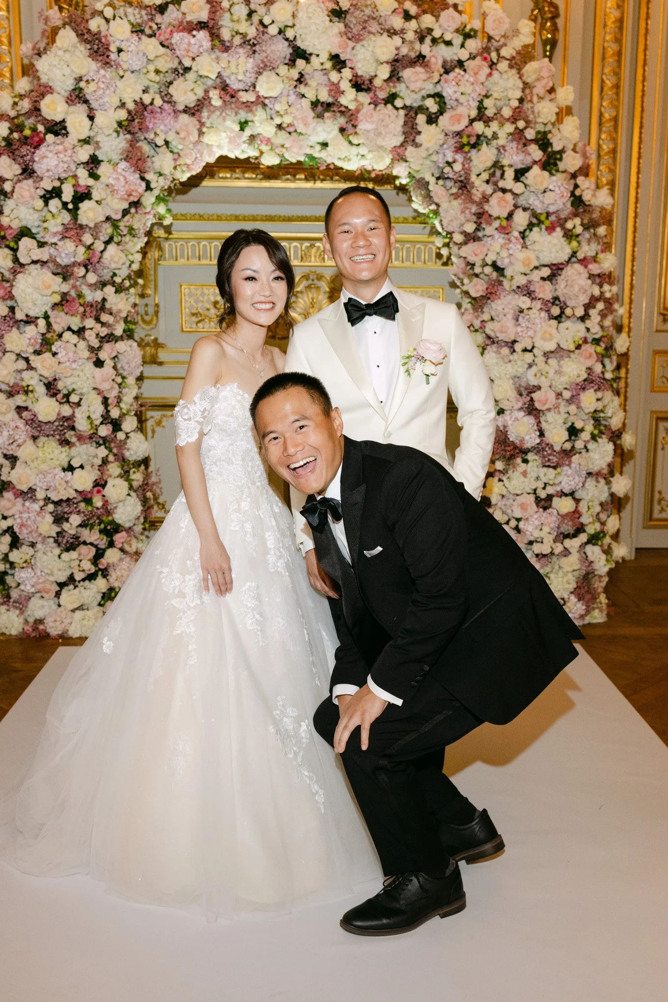A wedding party with a bride in a white dress, a groom in a tuxedo, and a man in a black tuxedo, standing in front of a floral arch with pink and white flowers.