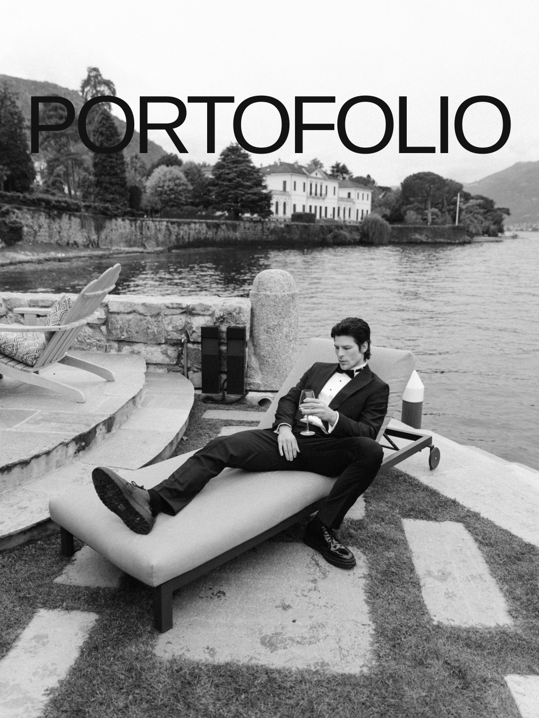 Man in tuxedo lounging on a pool chair by a waterfront, holding a glass of wine, with a house and trees in the background.