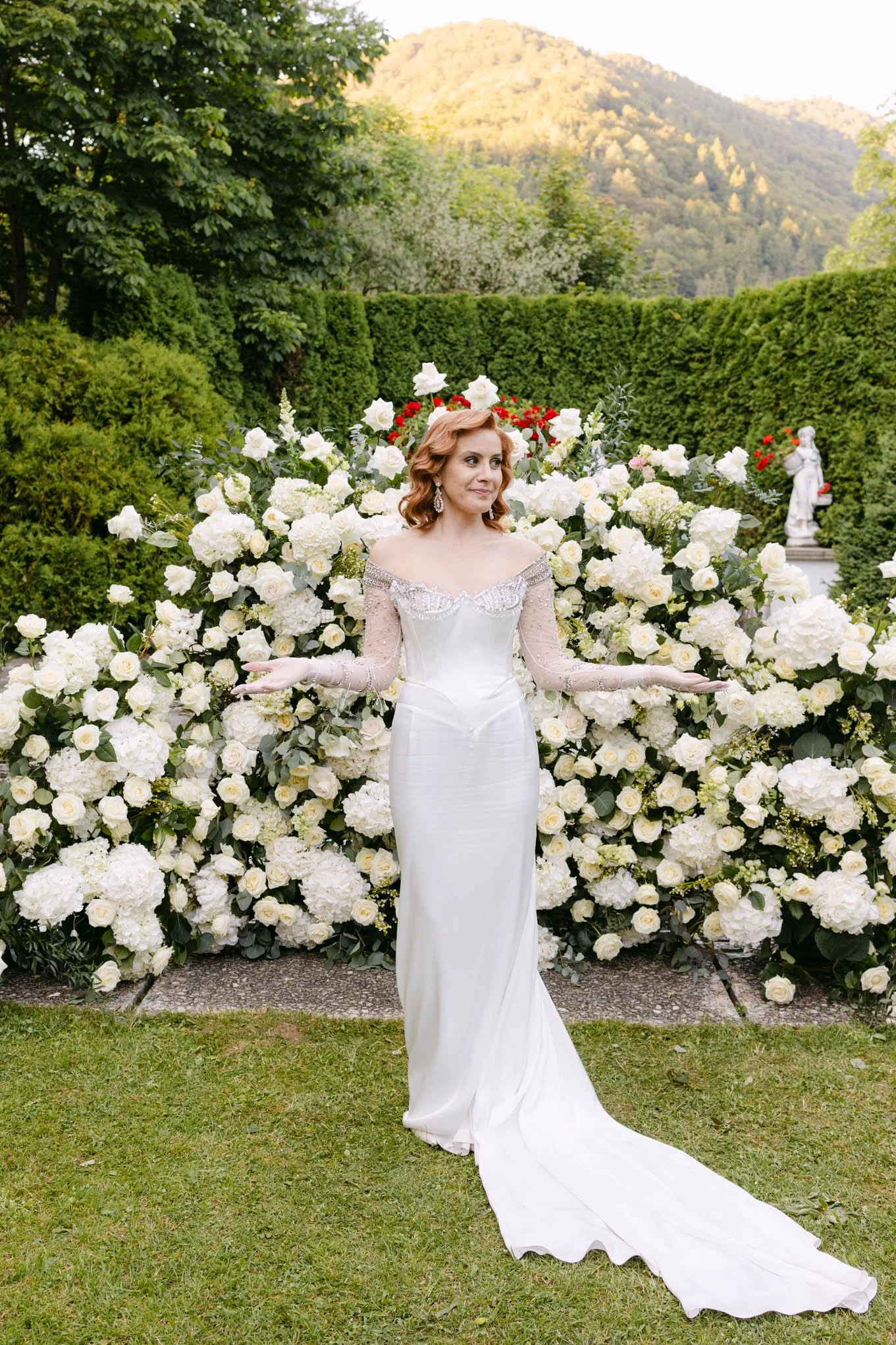 A bride in a white wedding gown stands outdoors in front of a floral backdrop of white roses and greenery, with a  Tuscany garden  the background.