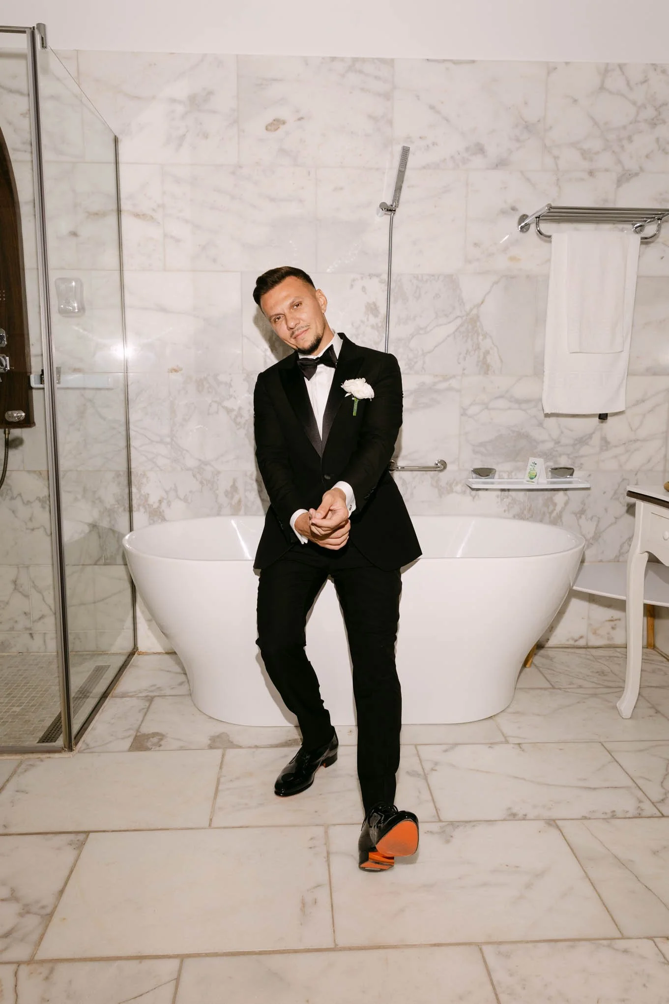 A man in a tuxedo with a white flower boutonniere, standing in front of a white bathtub. The background features marble walls and floor, a glass shower door, and a towel hanging on a rack. FAshion Vogue style groom!