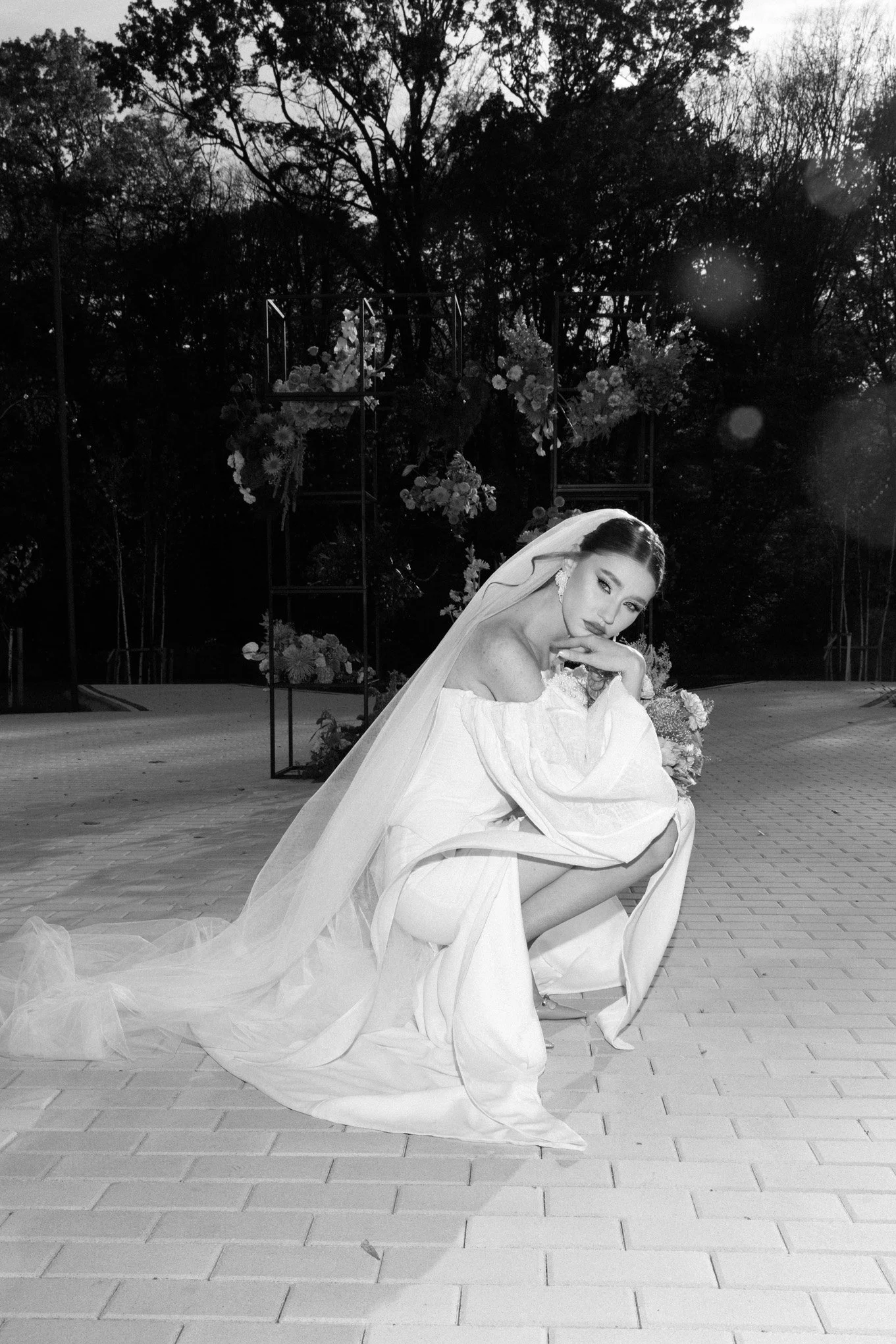 A woman wearing a wedding gown and veil, sitting on a brick surface outdoors, with a floral arch and trees in the background.