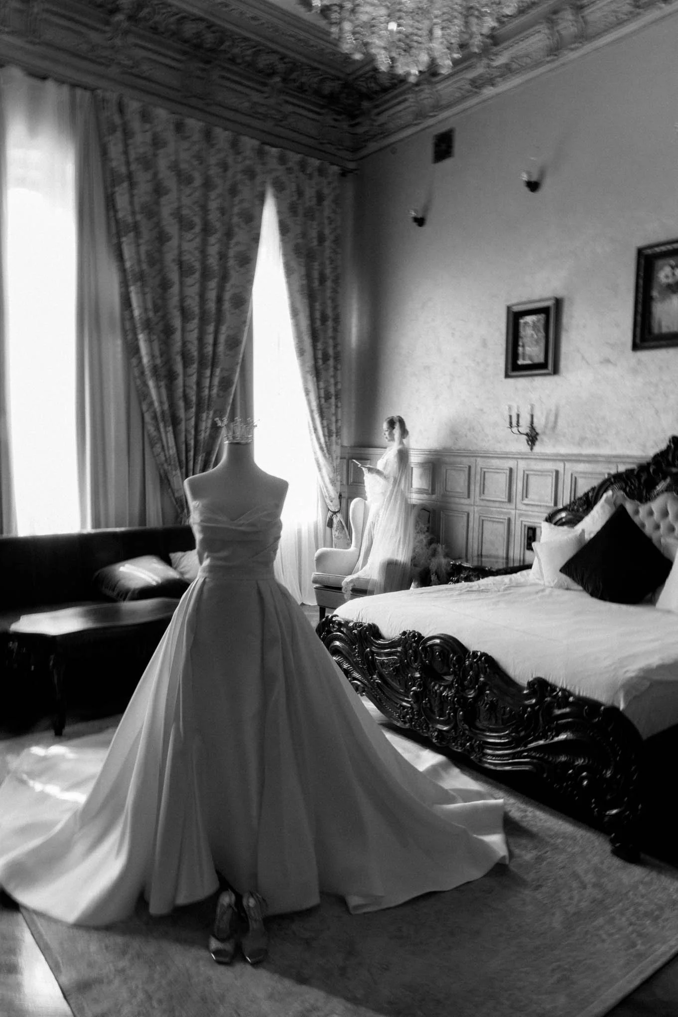 A luxurious bedroom with ornate furniture, a mannequin dressed in a wedding gown, and a woman in a wedding dress reading a book by the window. Italian style as seen in vogue weddingz, Elle weddings, Bazaar Weddings.