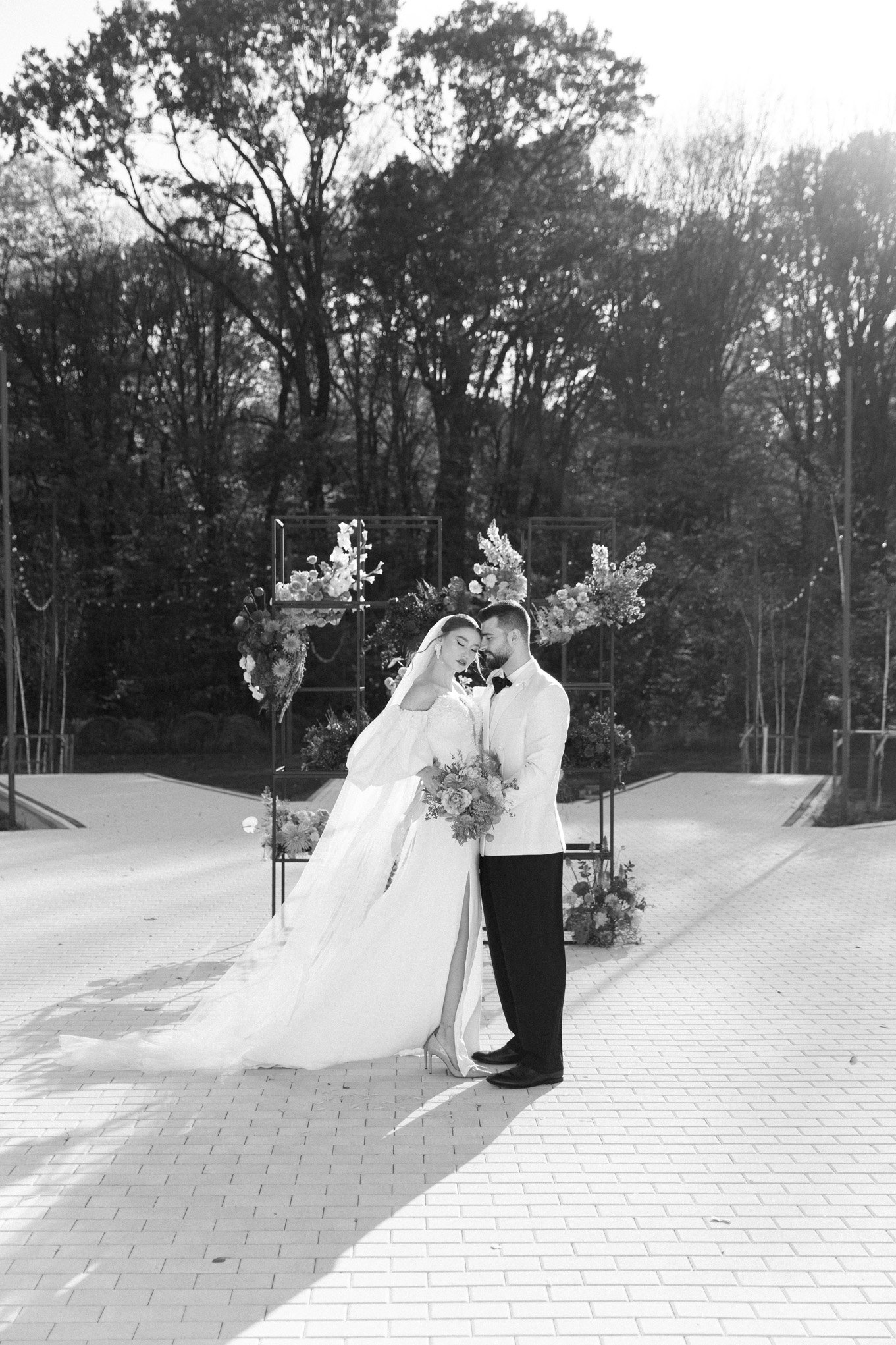 Black and white photo of a bride and groom standing close together outdoors, with the bride holding a bouquet and wearing a long veil and dress, and the groom in a tuxedo jacket and bow tie, in front of a flowered arch and trees.