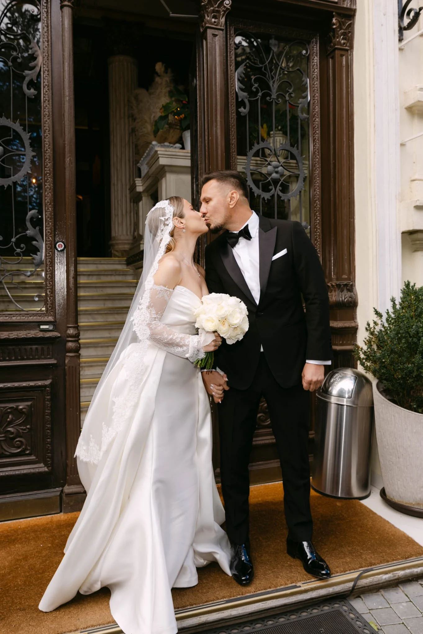A bride and groom kissing outside a building, with the bride holding a bouquet of white roses, dressed in a white wedding gown with lace sleeves, and the groom in a black tuxedo with a bow tie. Ass seen in Vogue!