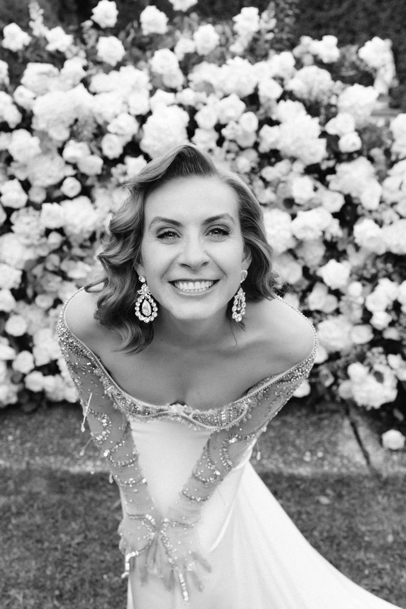 A woman in an elegant wedding dress with beaded embellishments and large earrings, smiling and looking up towards the camera, in front of a wall of lush flowers from a Tuscany style garden Villa.