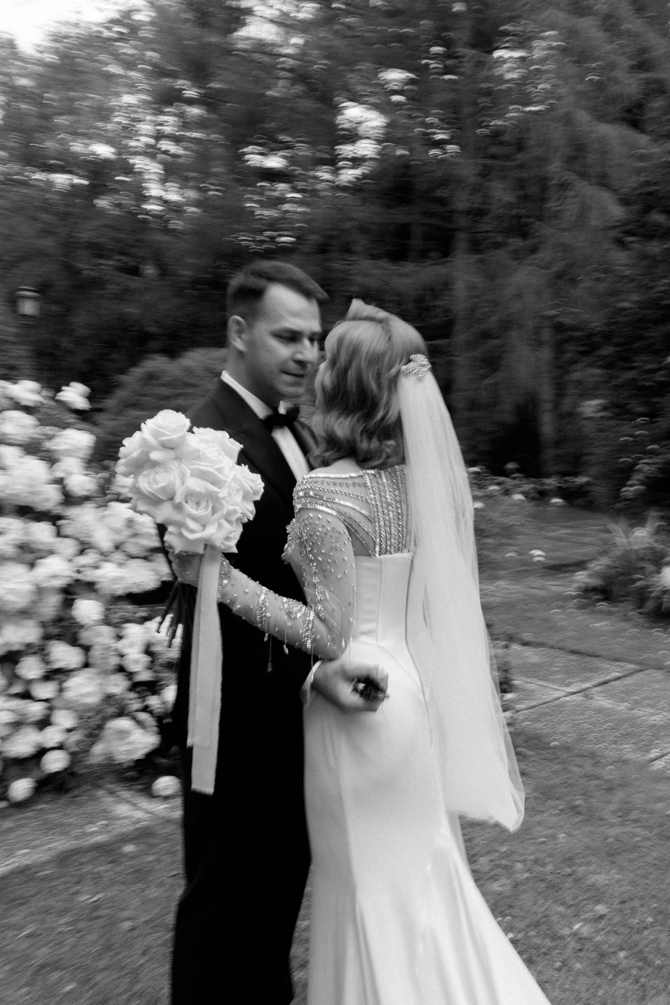 A black and white photo of a bride and groom embracing outdoors, with the bride holding a bouquet of roses, surrounded by trees and flowers.