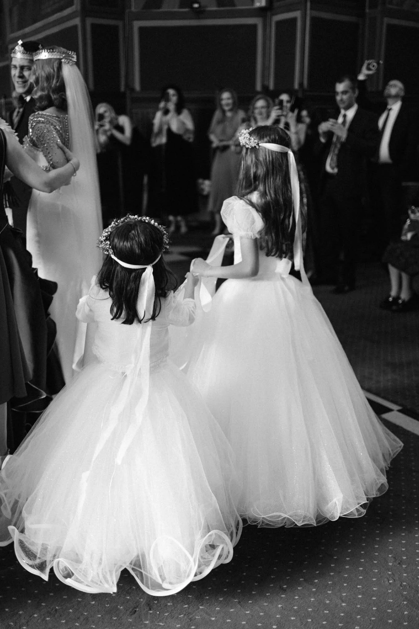 Two young girls in white princess dresses and floral headbands holding hands during a wedding ceremony, with guests watching and taking photos in the background. Italian style wedding.