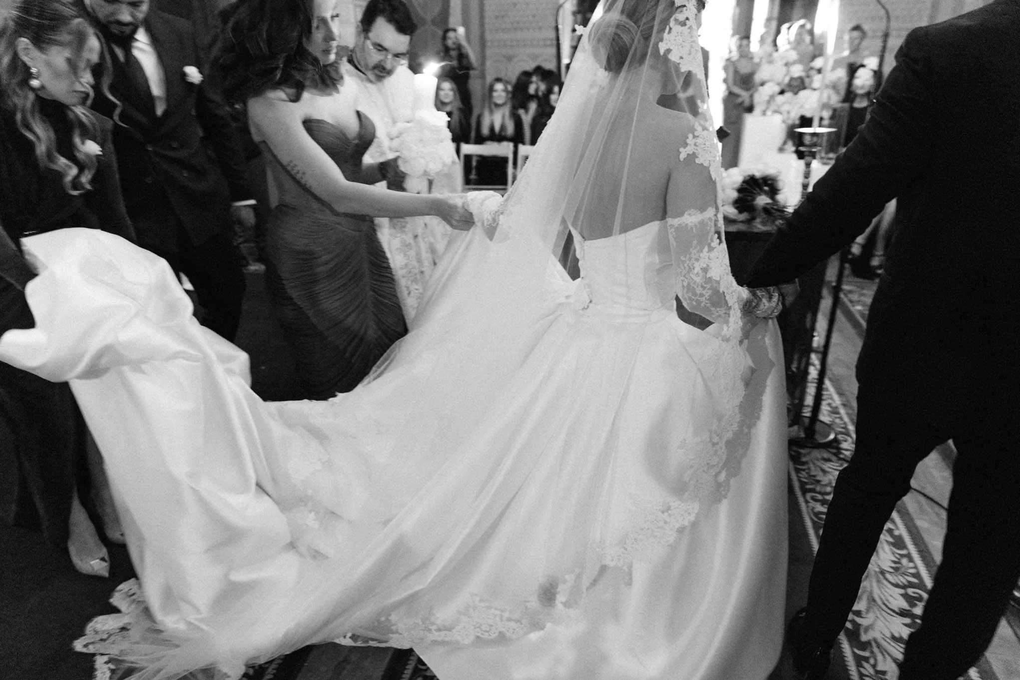 Wedding ceremony with bride in a white gown and veil, holding hands with groom, surrounded by guests indoors. BW shot of french style wedding, as seen in south of france weddings!