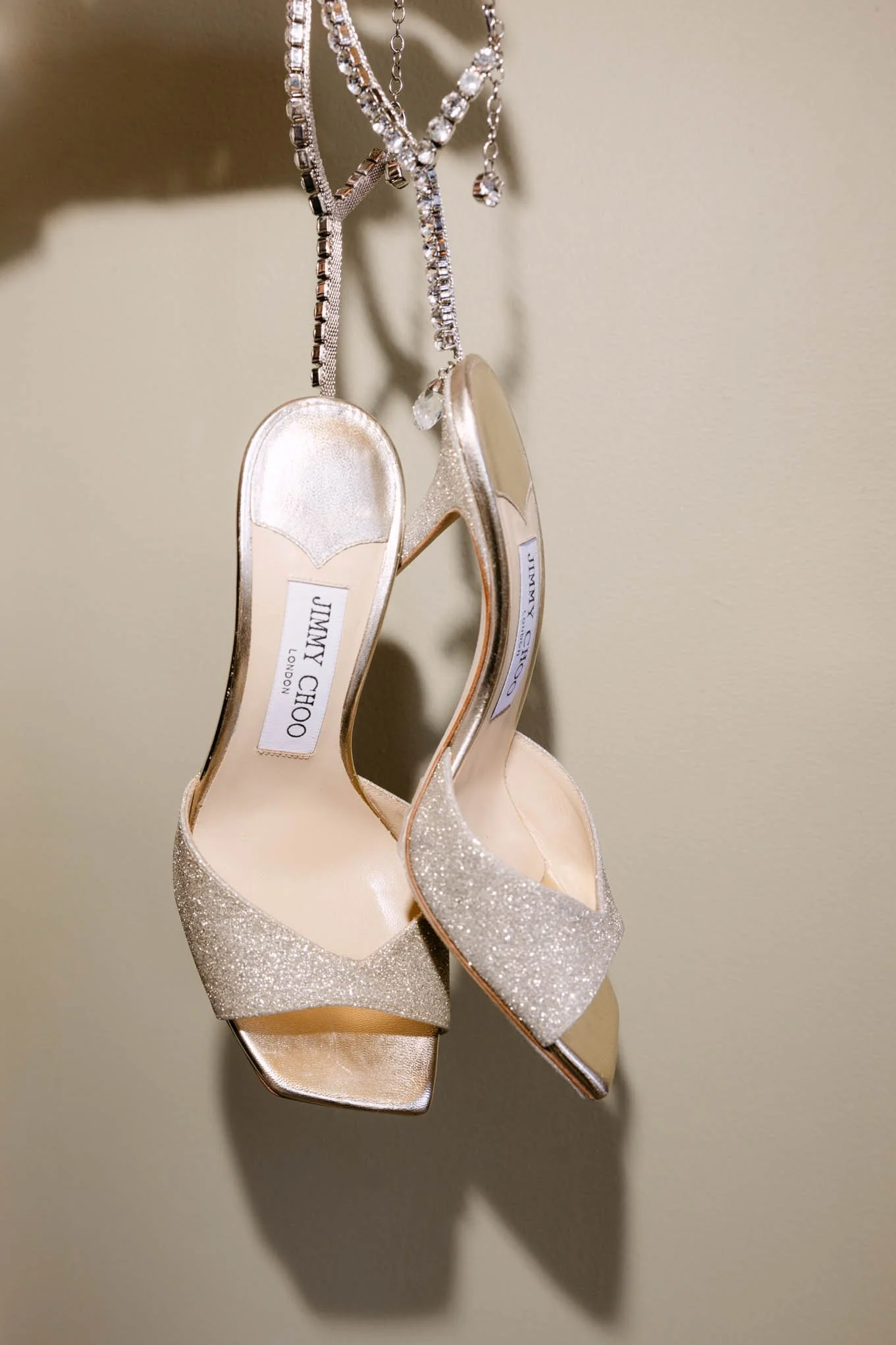 A pair of glittery gold Jimmy Choo high-heeled sandals with open toes, hanging from a jewelry chain and shadowed on a plain wall.Vogue style!