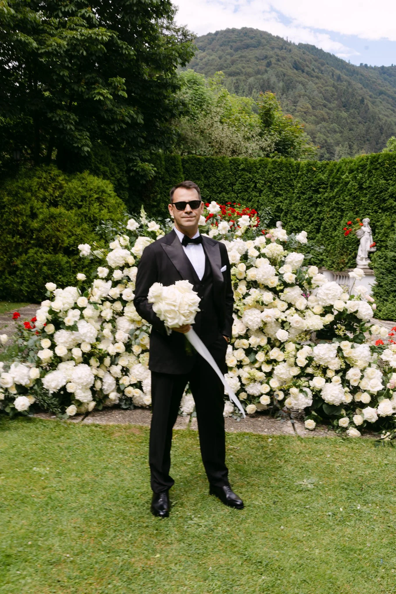 A man in a tuxedo and sunglasses holding a bouquet of white roses, standing in front of a garden with white and red flowers and green hedges, with an italian style garden.
