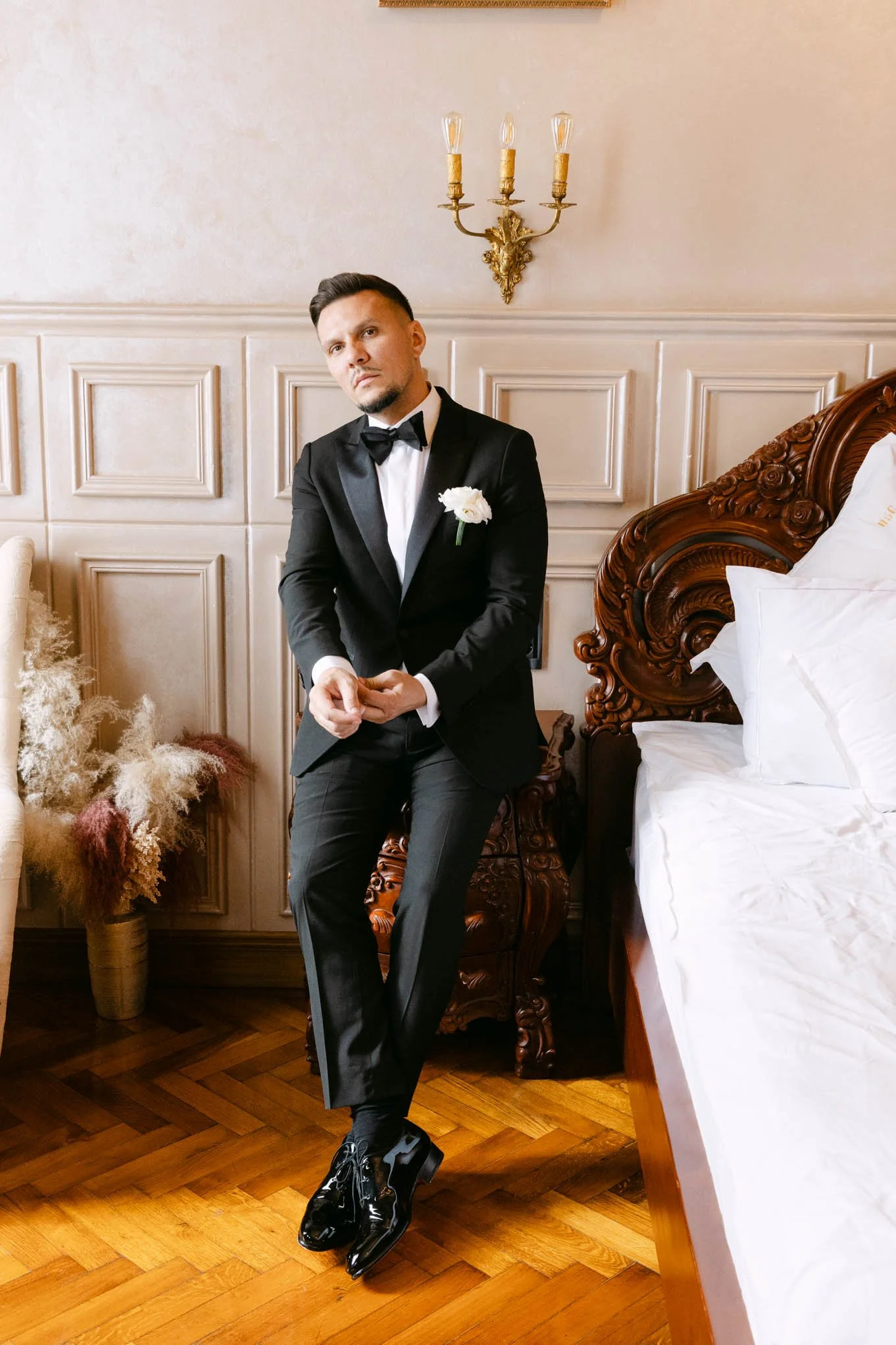A man in a tuxedo with a white flower boutonniere, standing beside a bed in a luxurious room, wearing shiny black dress shoes. Fashion groom as seen in vogue!