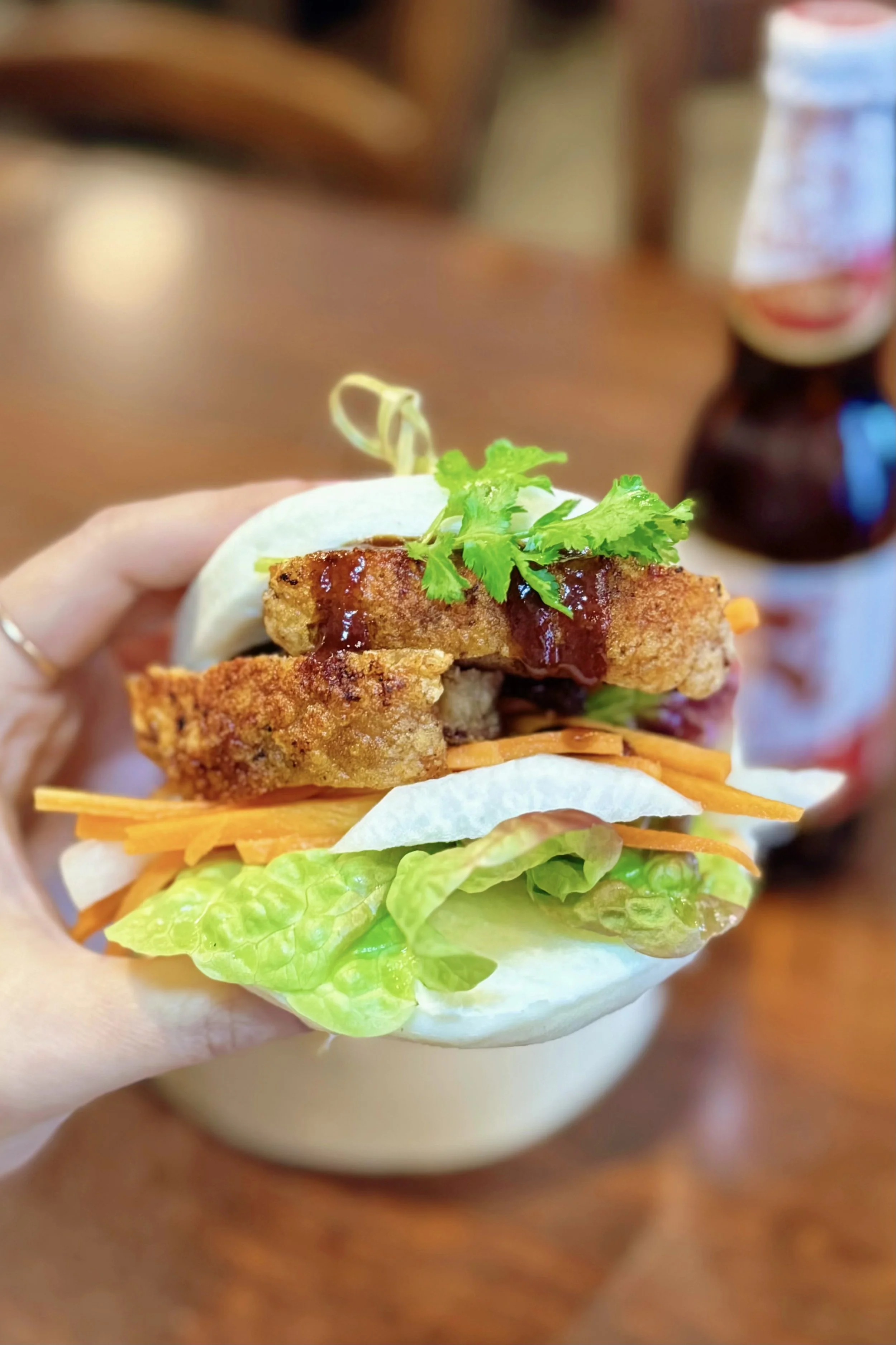Close-up of a person holding a burger with lettuce, shredded carrots, fried chicken fillet, cilantro, and a drizzle of sauce in a bun.