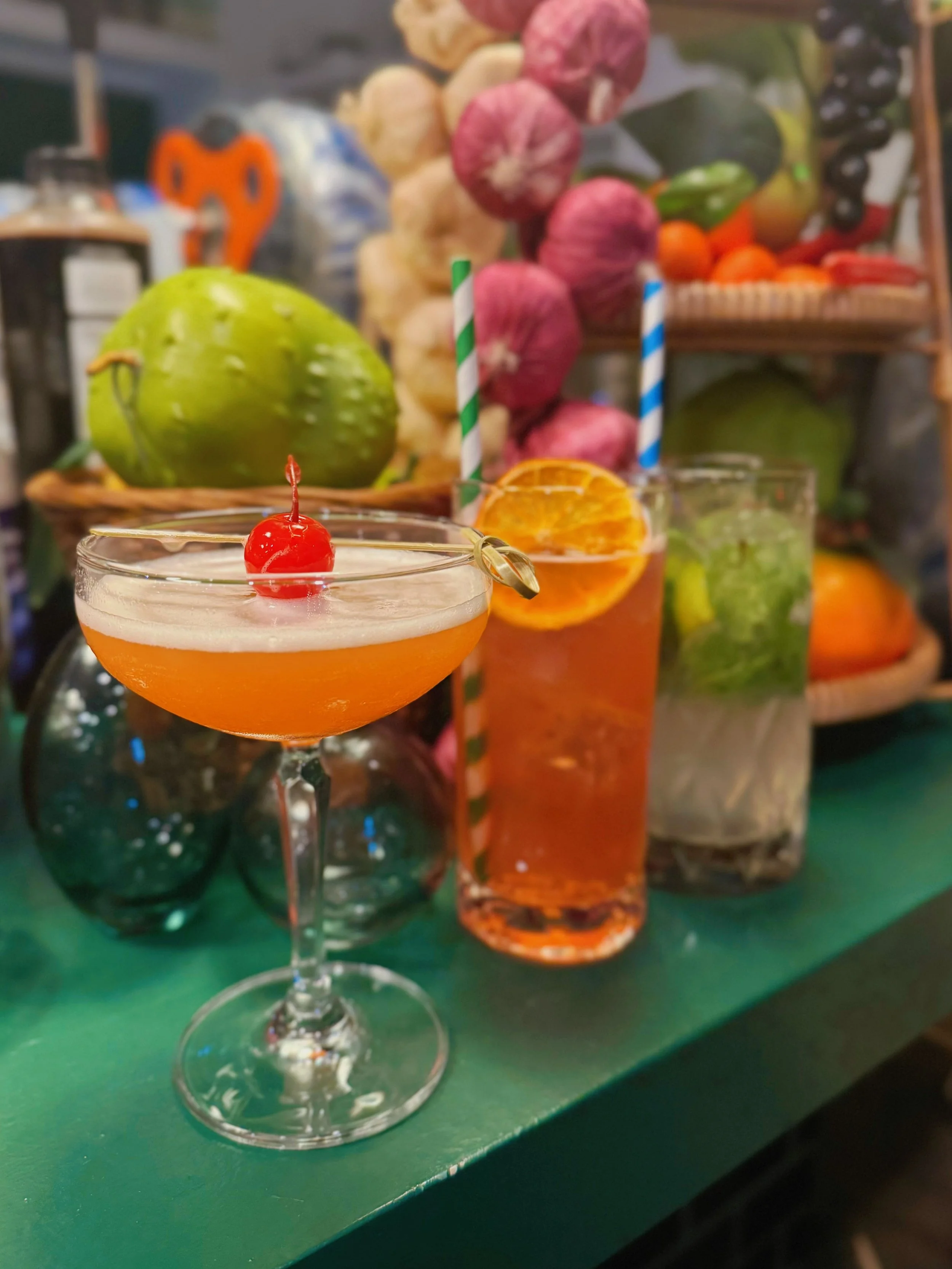 Several colorful cocktails with fruit garnishes and striped straws on a green table, with a background of fruits and vegetables.