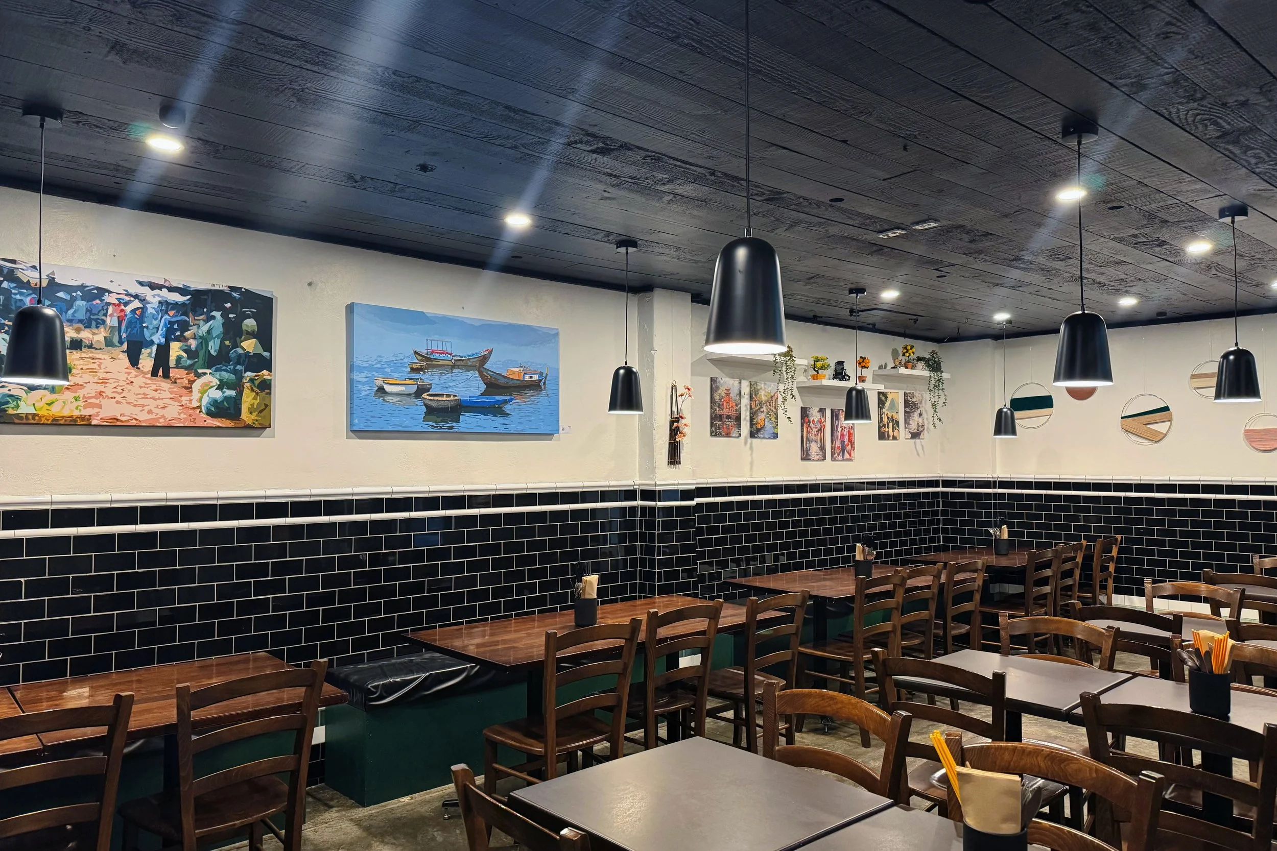 Interior of a restaurant with wooden tables and chairs, black pendant lights hanging from a dark wood ceiling, and artwork on the white walls depicting various scenes including boats and outdoor settings.