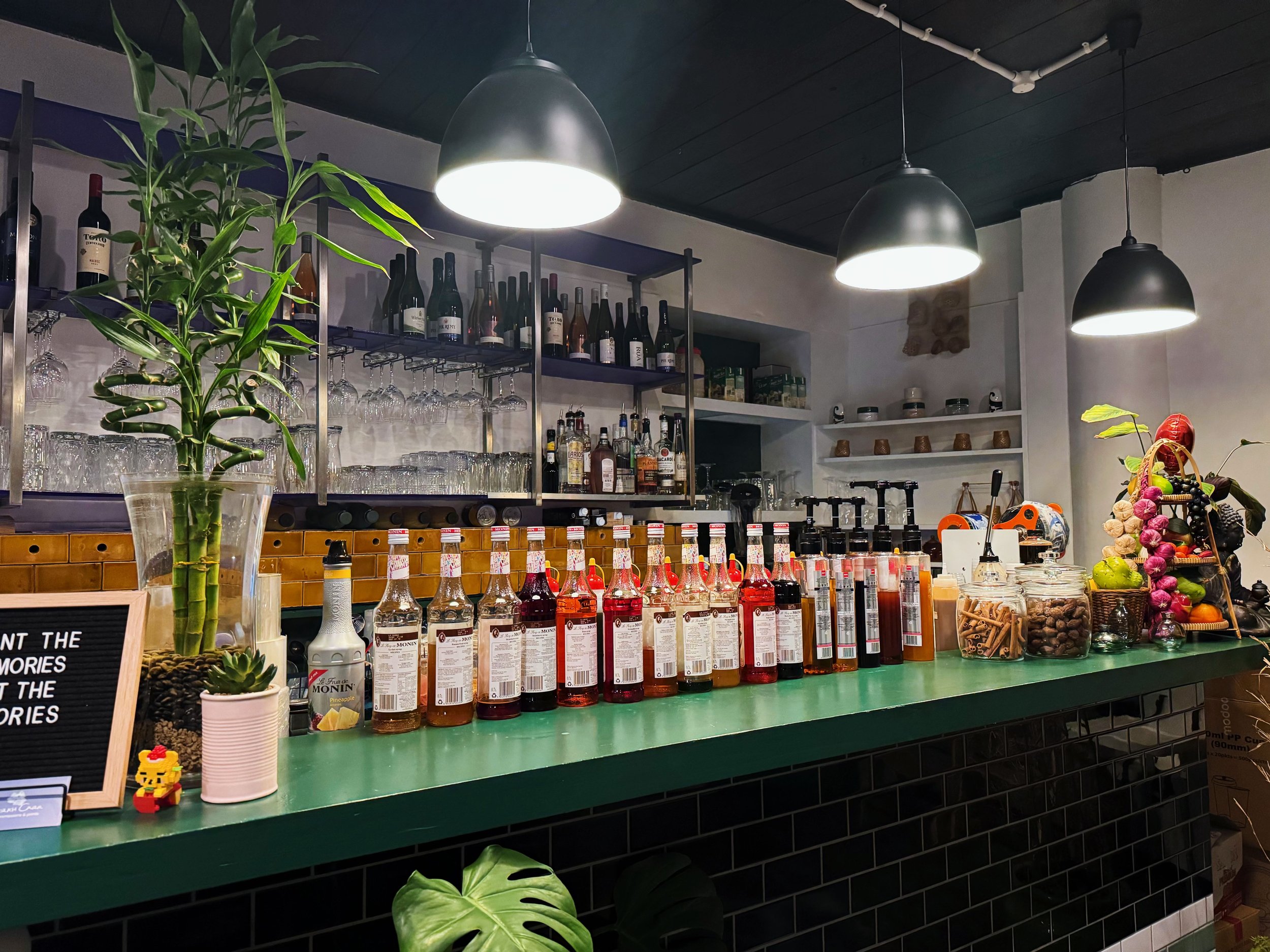 A bar or cafe counter with a green countertop displaying bottles of Monin syrup, a potted bamboo plant, and a small lego figure. In the background, there are shelves with wine bottles and glasses, along with other bottles and kitchenware. Overhead, t