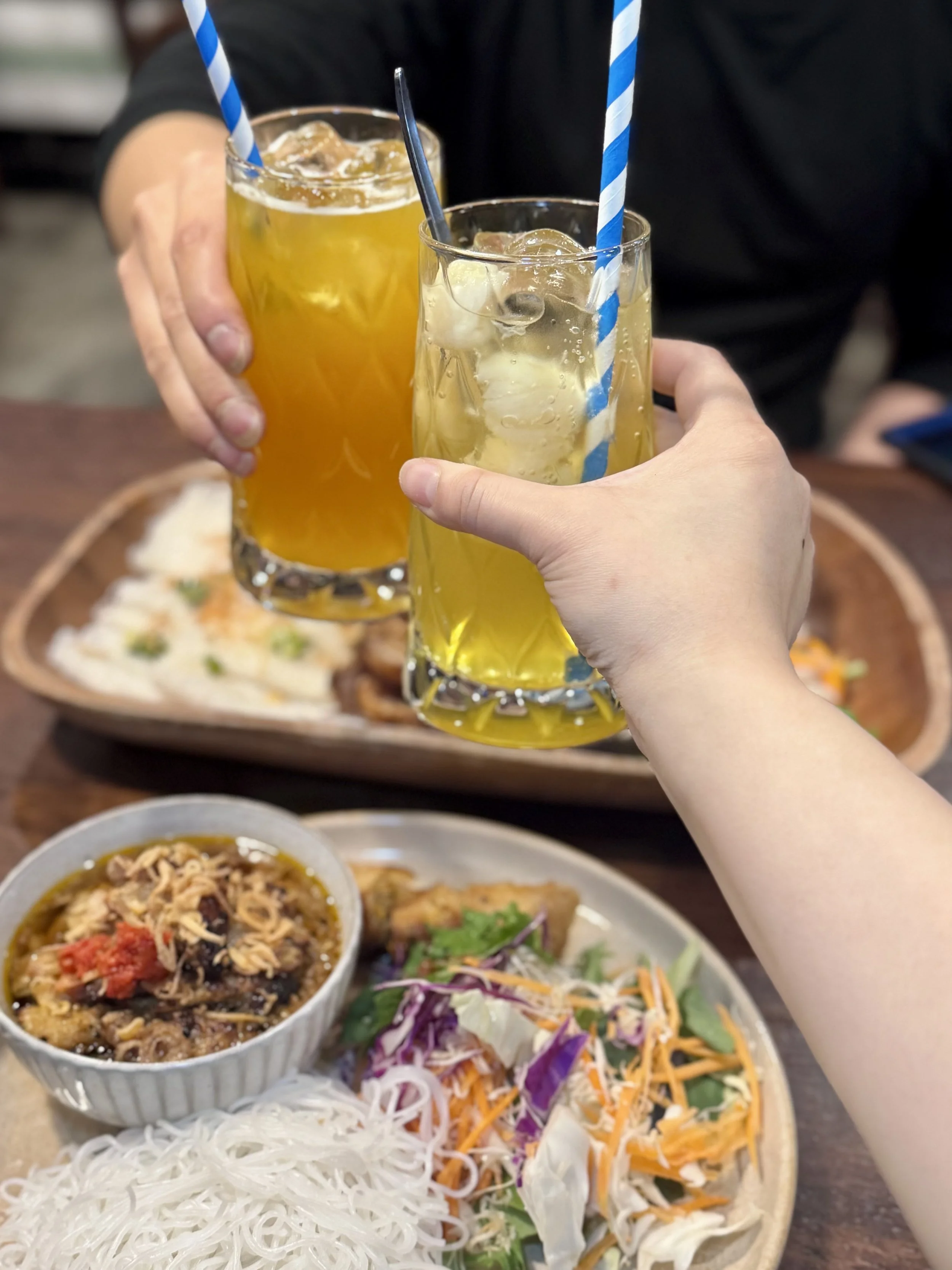 Two people clinking glasses with yellow beverages and straws in a dining setting. In the background, there is an assortment of Asian dishes including rice noodles, a bowl of stir fry, and vegetable salad on a wooden table.