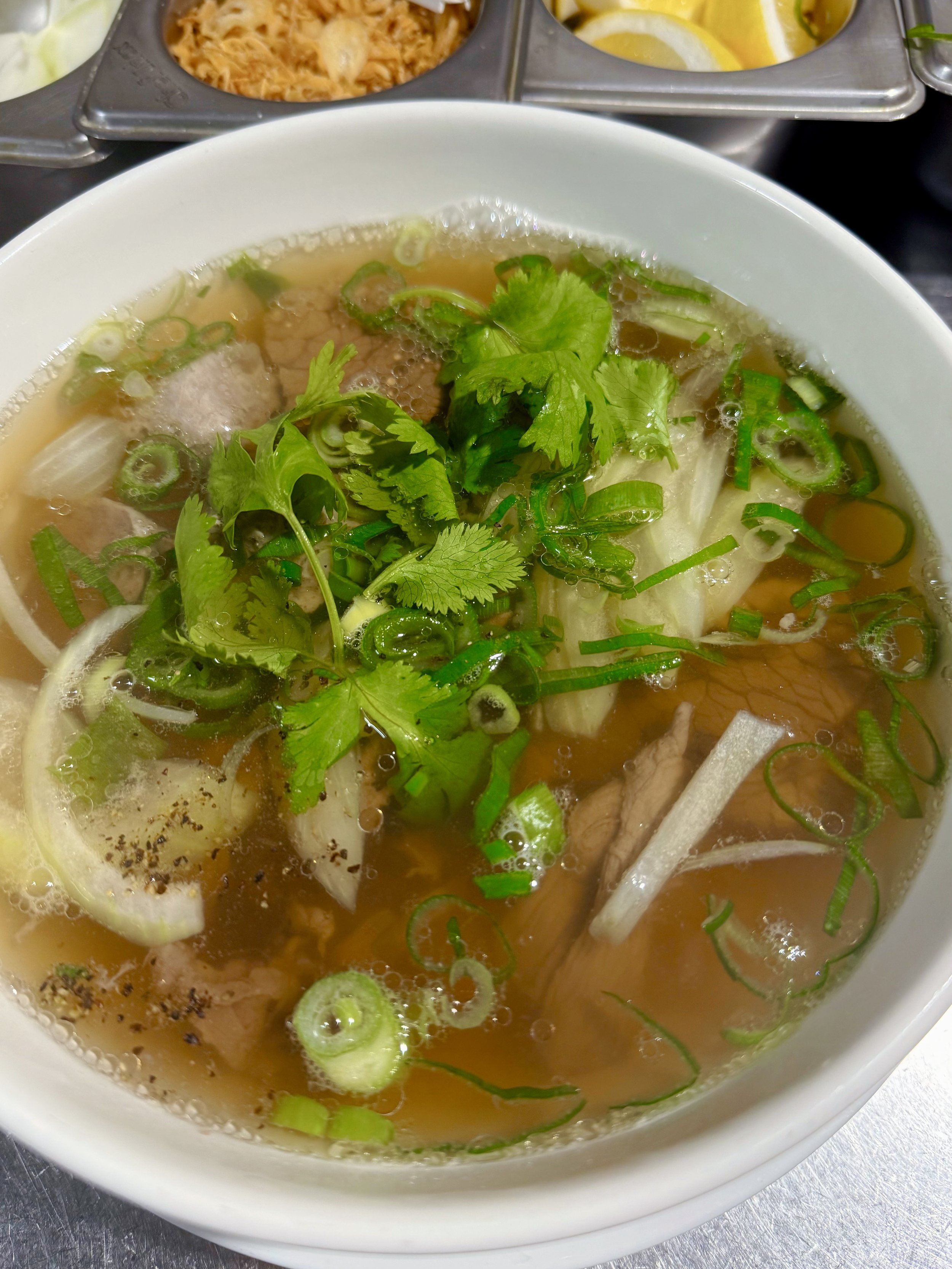 A bowl of hot soup with green onions, cilantro, and slices of onion and meat. There are containers of lemon slices and shredded cheese in the background.