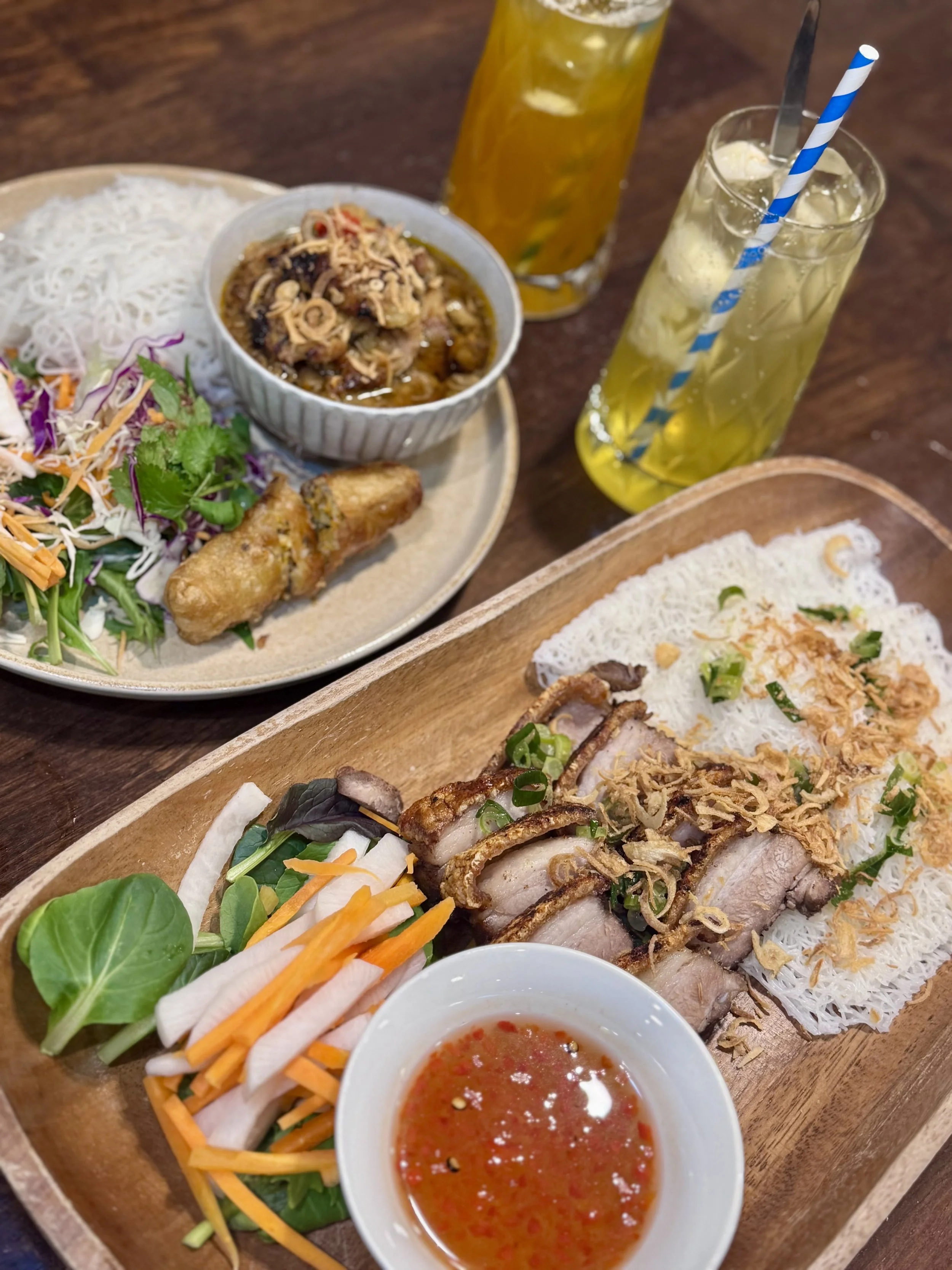 Assorted Vietnamese dishes including rice paper spring rolls with vegetables, sliced pork, fried spring roll, fresh salad, a bowl of Vietnamese grilled pork with noodles, two glasses of yellow-colored drink with ice, and a small bowl of dipping sauce