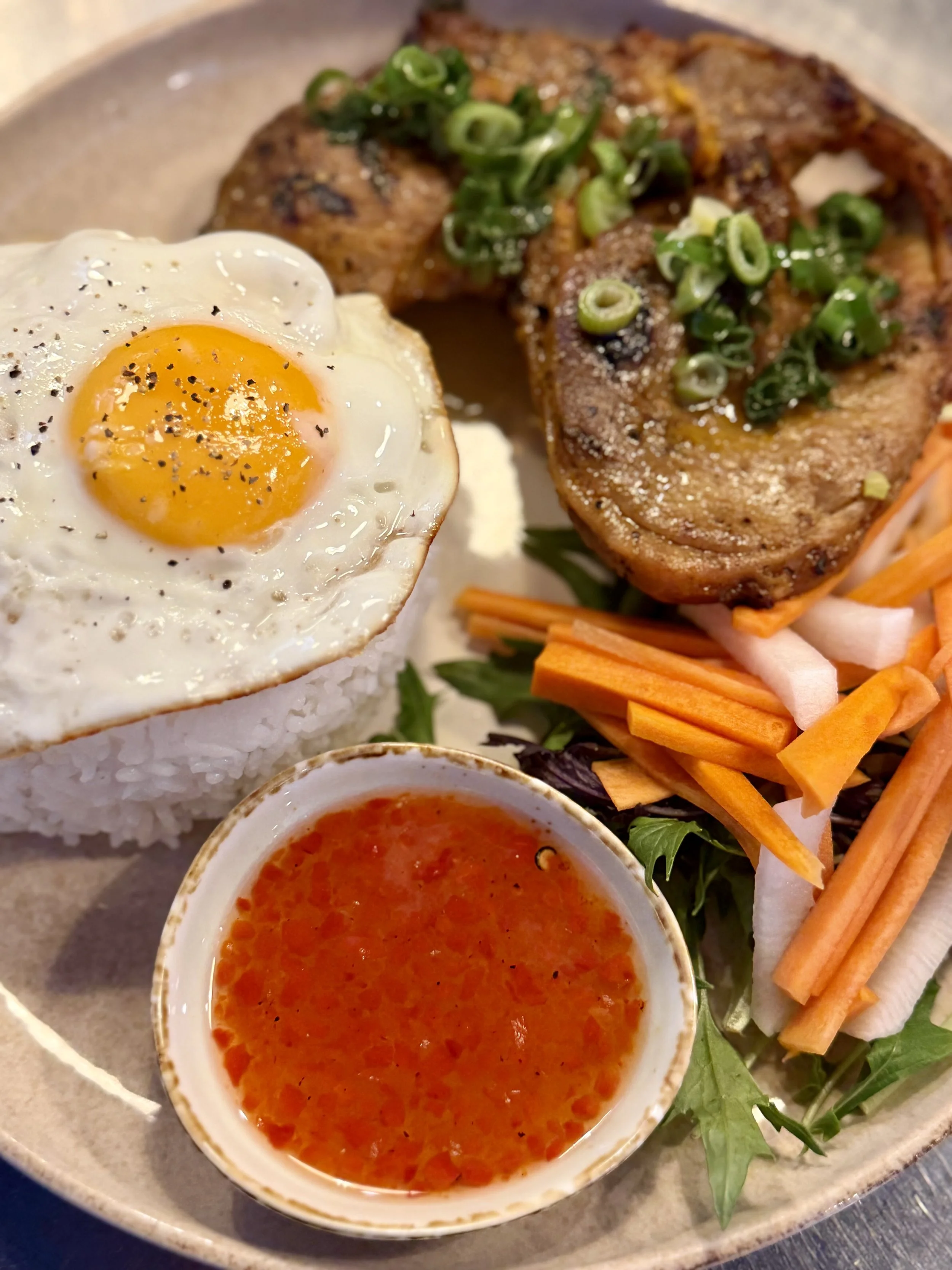 A plate of Vietnamese-style grilled pork chop, served with a fried egg, pickled vegetables, and a small bowl of dipping sauce.