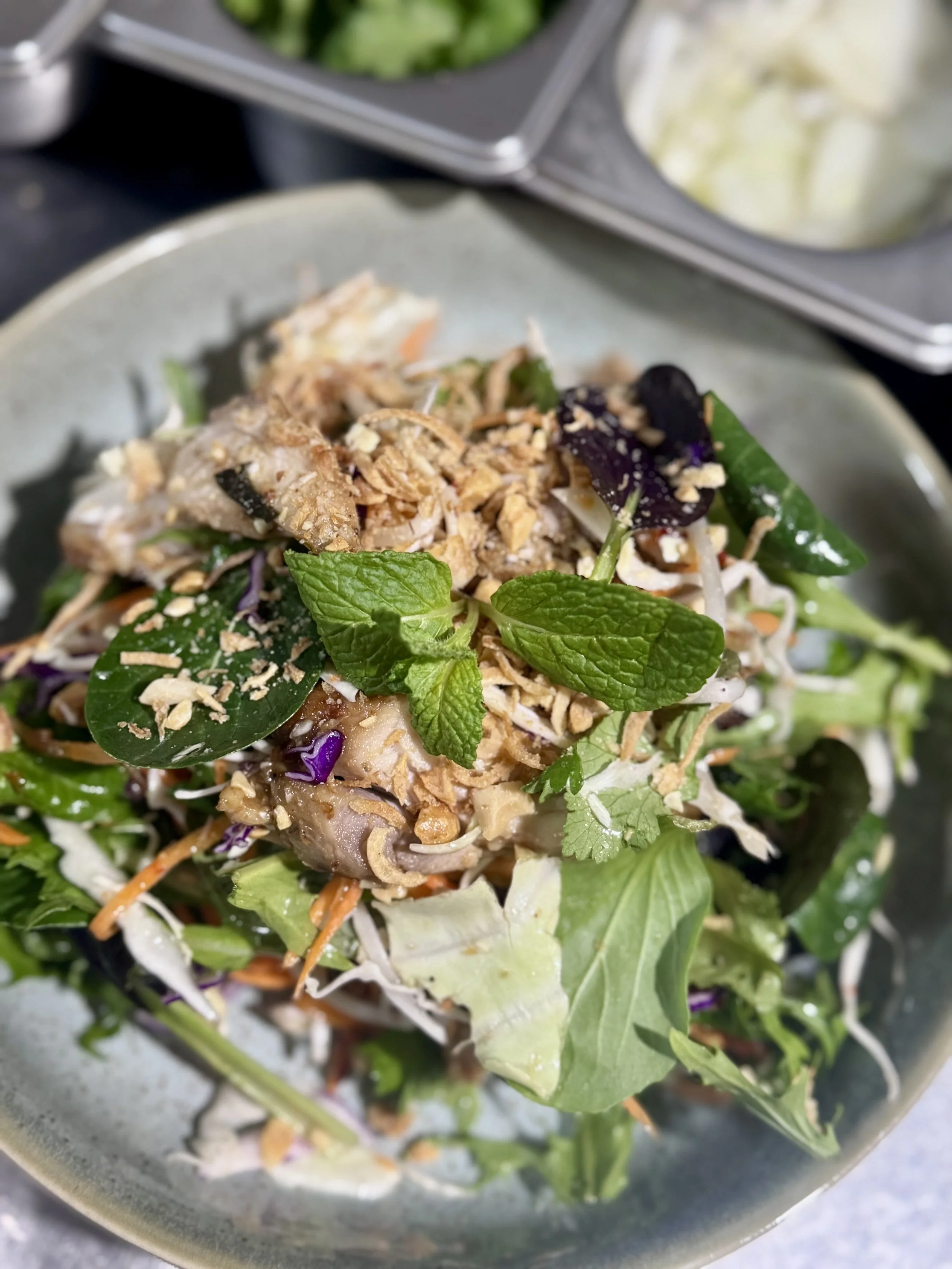 A salad with mixed greens, shredded vegetables, herbs, chopped nuts, and shredded cheese in a gray bowl.