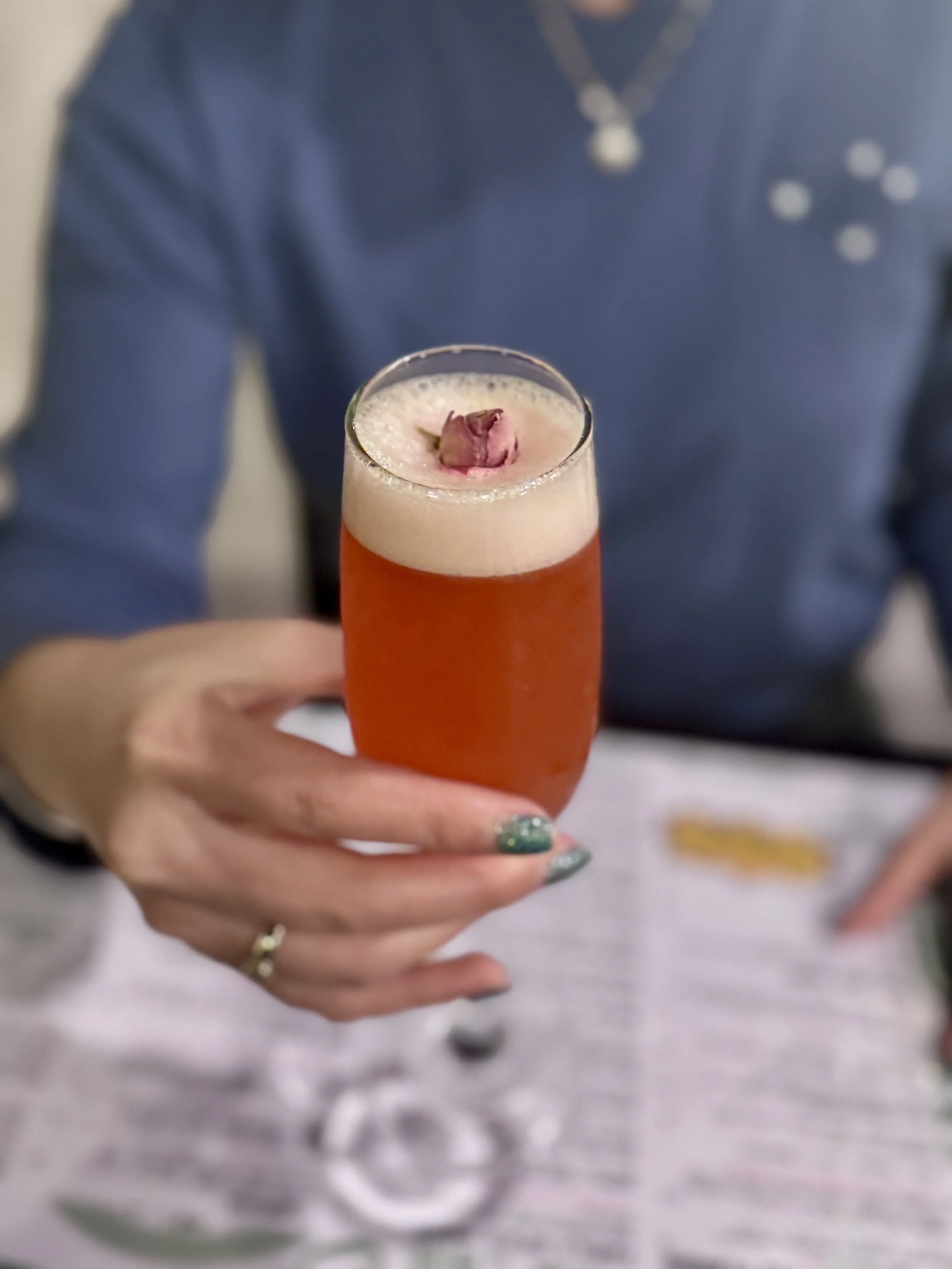 A person holding a tall glass of pinkish-orange beverage garnished with a pink flower petal.