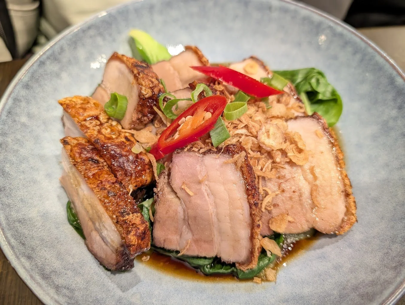 Dish with slices of roasted pork, garnished with chopped green onions, sliced red chili peppers, fried shallots, and greens, served on a ceramic plate.
