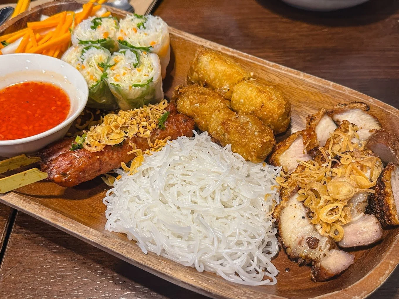 Assorted Asian grilled meats, rice noodles, spring rolls, fresh vegetables, and dipping sauce on a wooden serving platter.