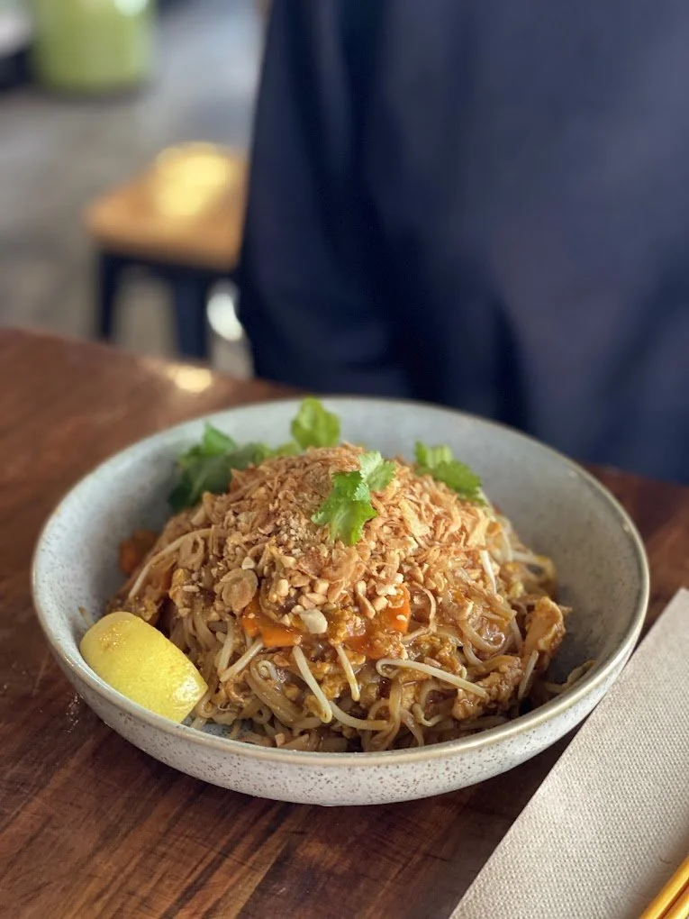 A bowl of Asian noodle salad garnished with chopped peanuts, cilantro, and a lemon wedge on a wooden table.