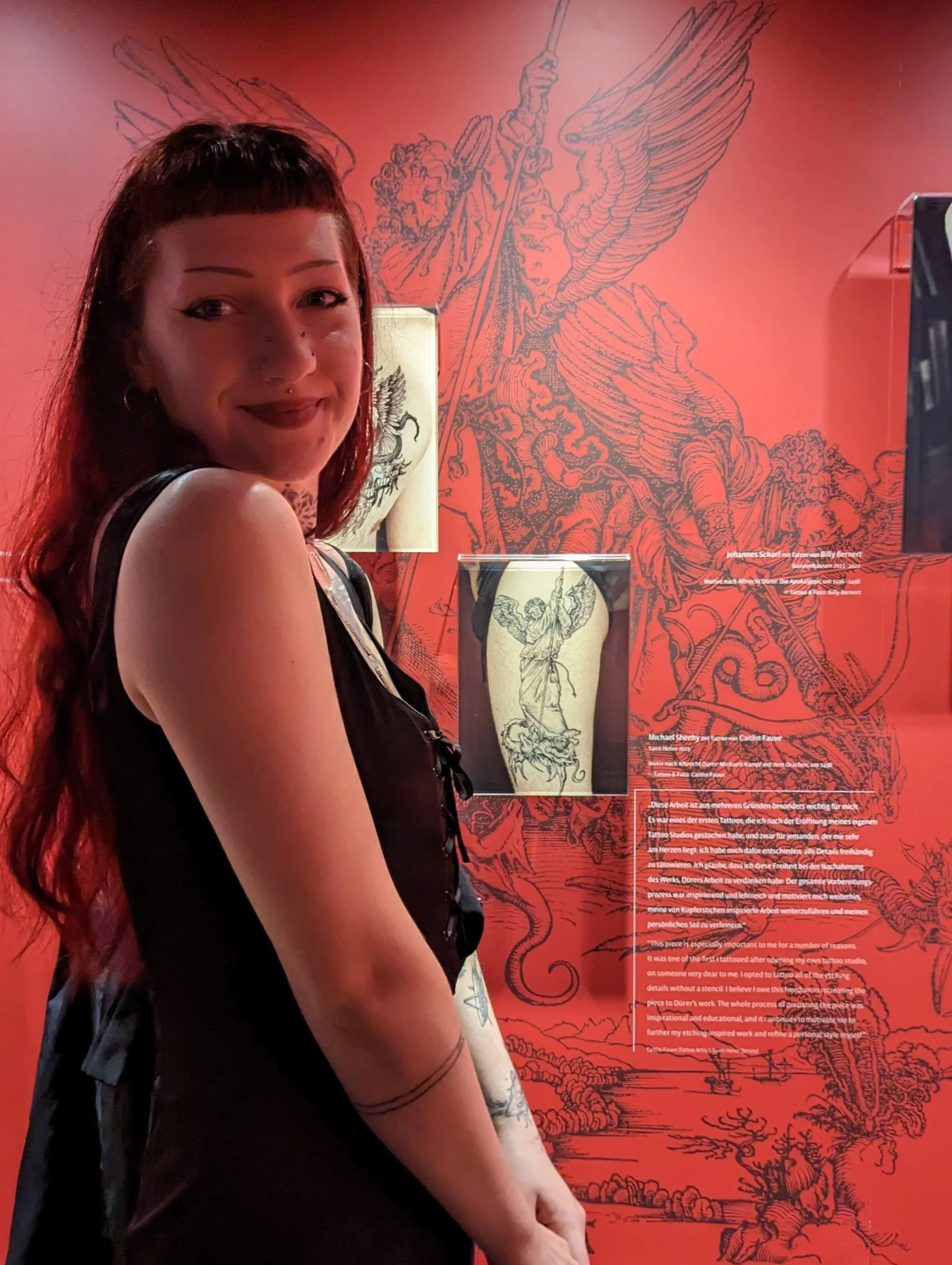 Me standing side on smiling, in front of a red wall showcasing a photograoh of one of my tattoos featured in at the Albrecht Durer House, Nuremburg, 2024.