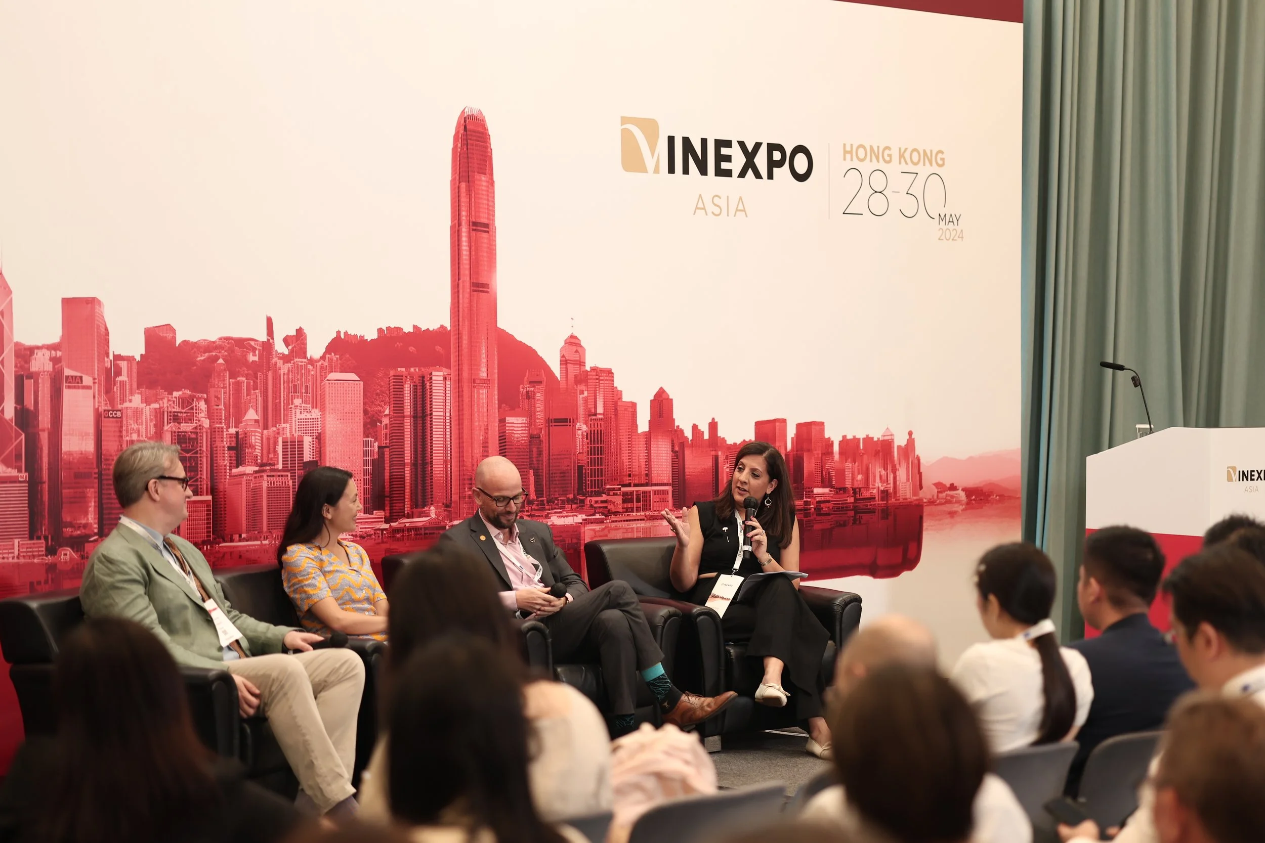 A panel discussion at a conference with four people sitting on a black couch on stage, one woman speaking into a microphone and gesturing. Audience members are seated in front, and a large backdrop features Hong Kong's skyline with the event info: MI