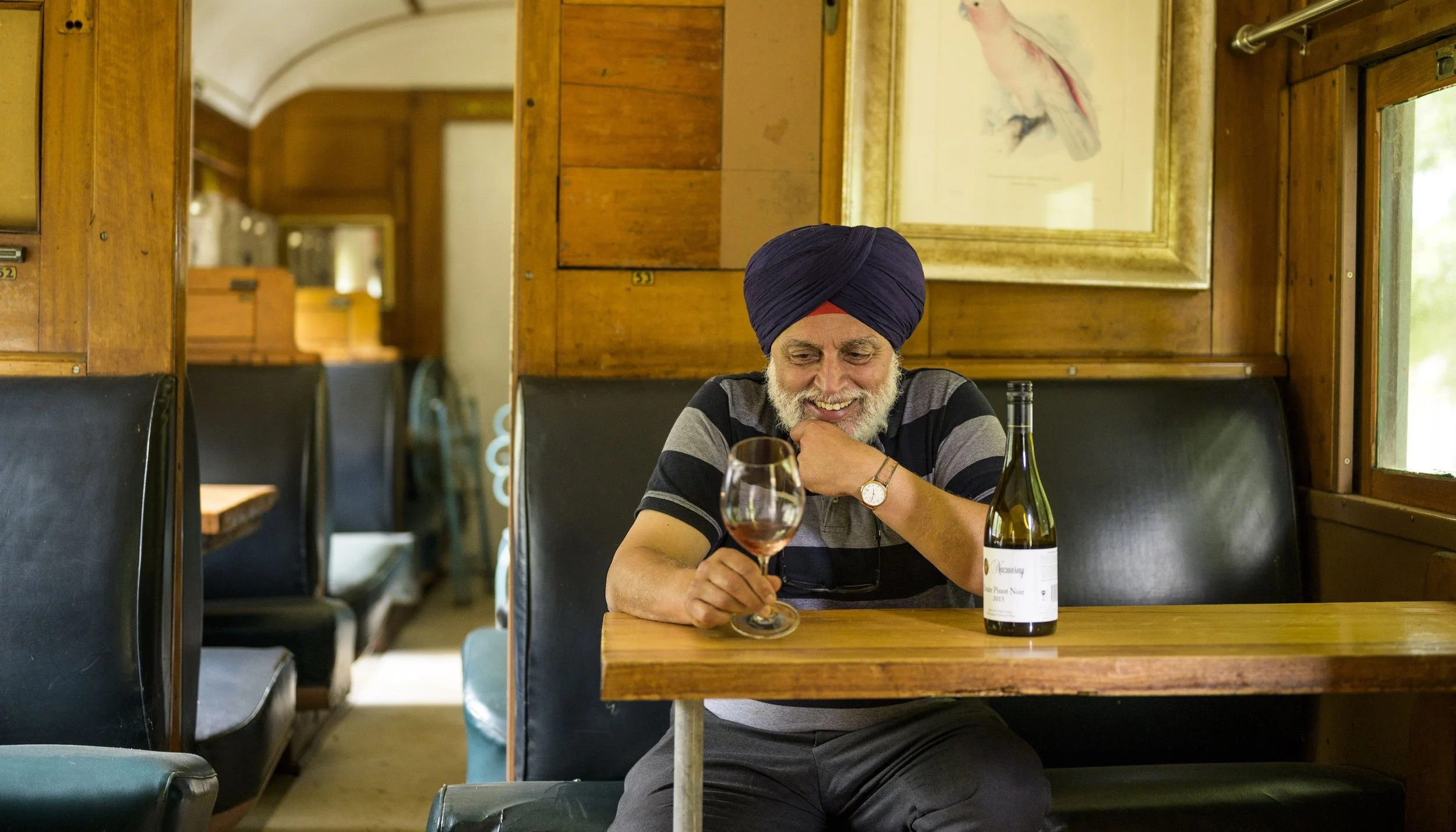 An elderly man with a white beard and a purple turban sits at a wooden table in a restaurant, smiling and holding a glass of red wine. There is a bottle of wine on the table, and the background features wooden paneling, a framed picture of a bird, and a window.