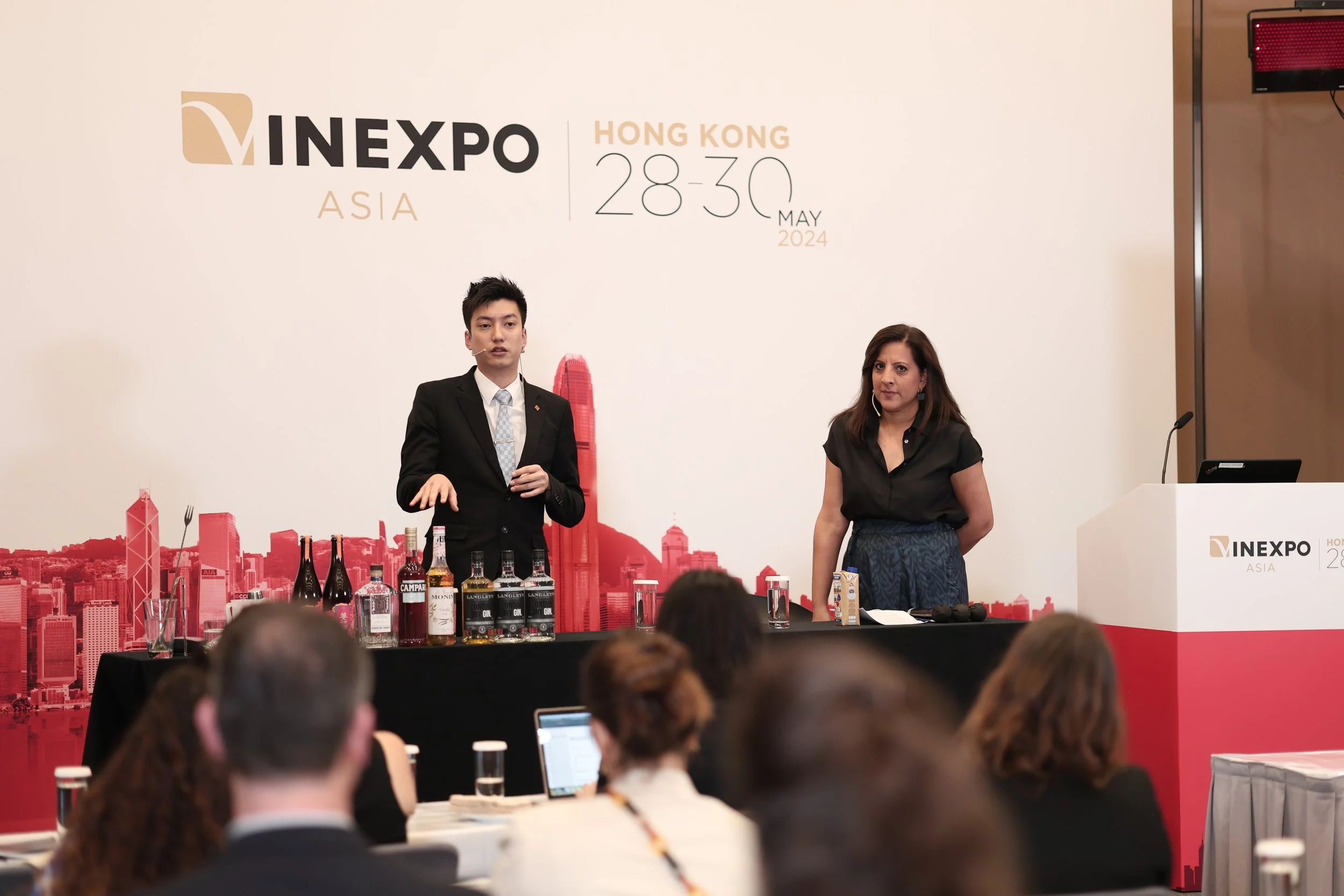 Nimmi Malhotra presenting at Vinexpo. She is standing behind a table with various bottles, at the VINEXPO Asia conference in Hong Kong, May 28-30, 2024, with the audience sitting in front.