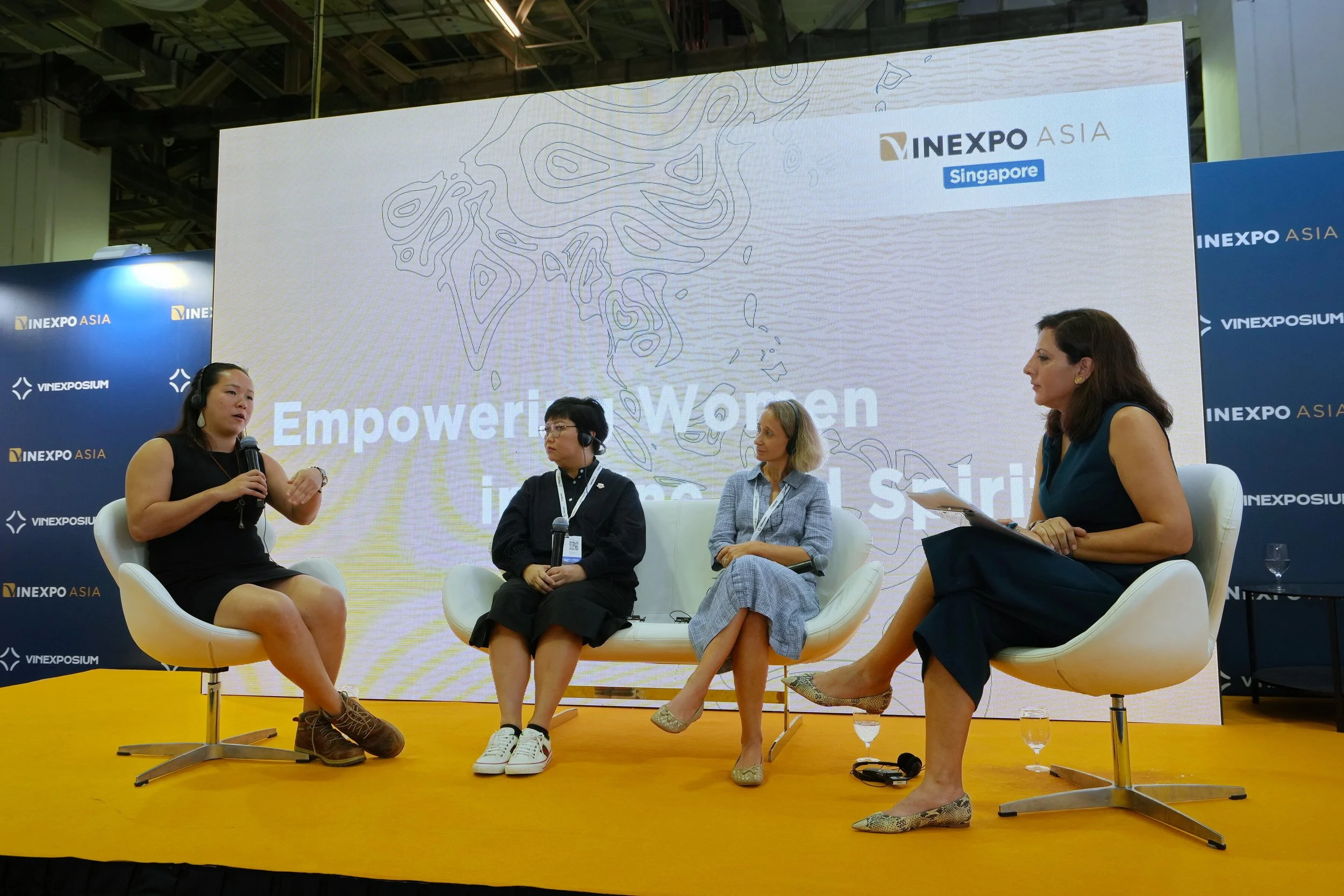 Four women, including Nimmi Malhotra seated on a stage at a panel discussion about empowering women, with a large screen behind them displaying 'Empowering Women in Asia' and 'INEXPO ASIA Singapore'. 