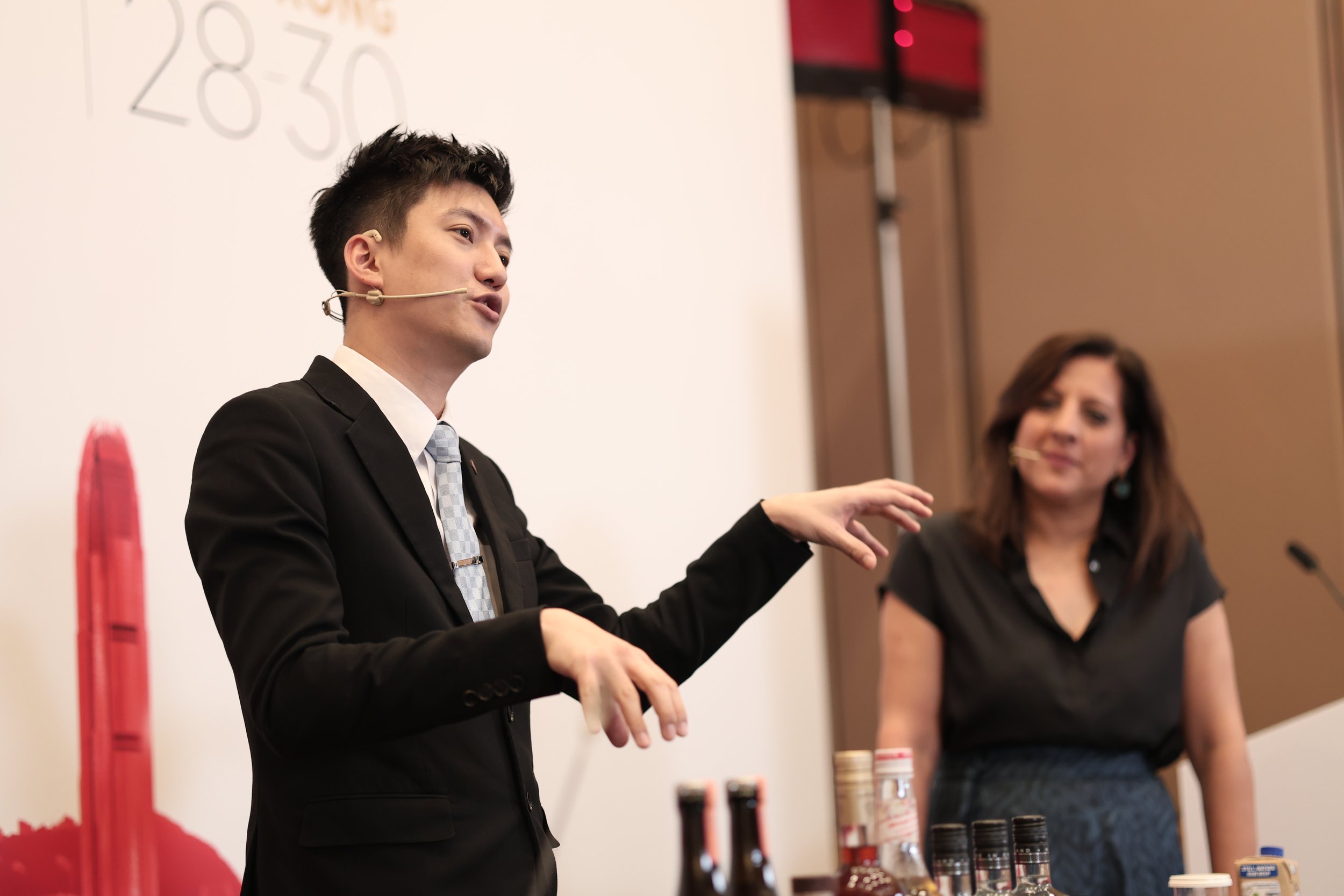 A man in a black suit with a microphone headset speaking at a presentation, with a woman standing beside him. There are bottles of condiments and some paper cups on the table in front of them.