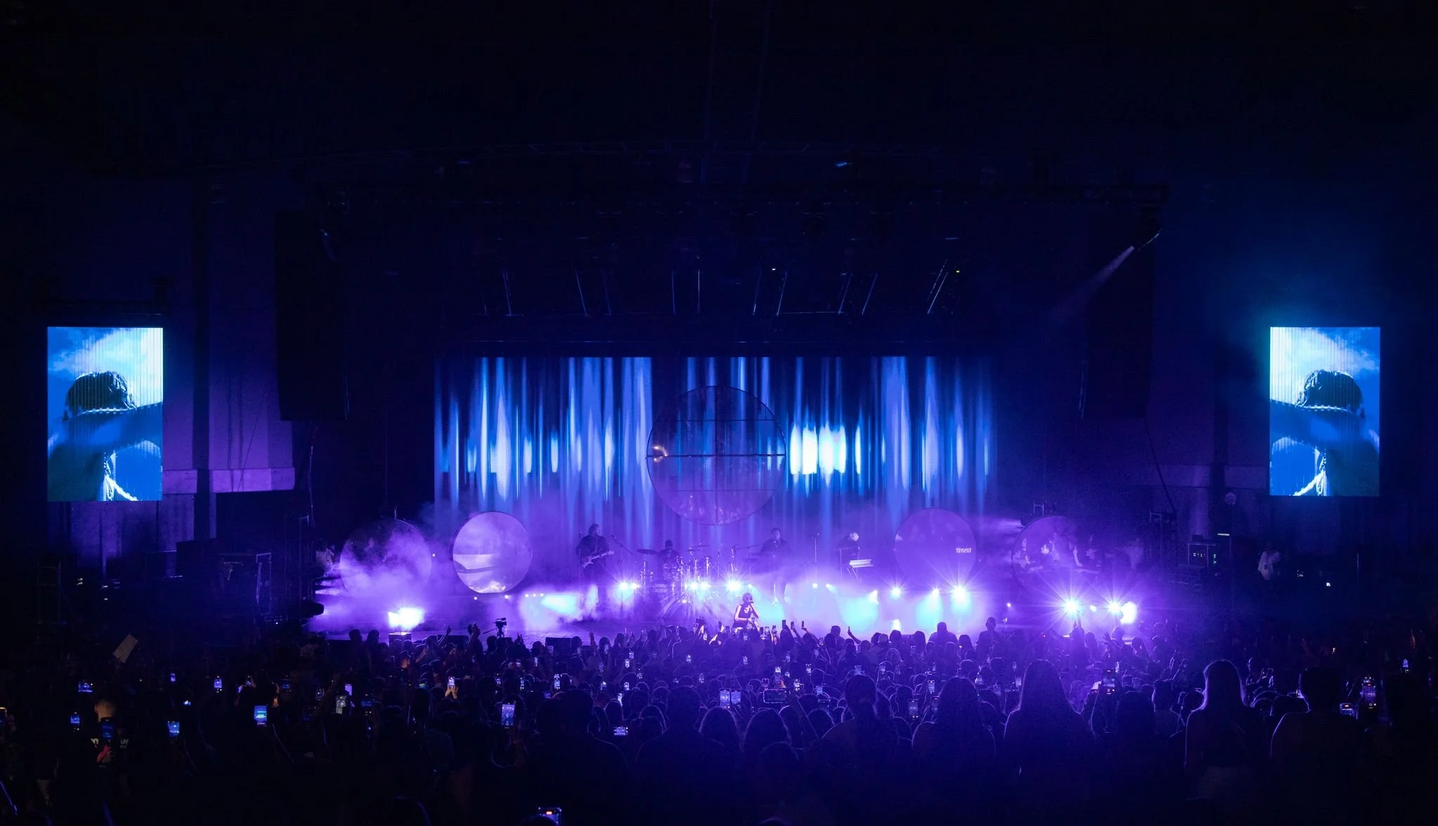 Concert stage lit with purple and blue lights, large screens showing a person with their arm over their face, audience with phones raised.