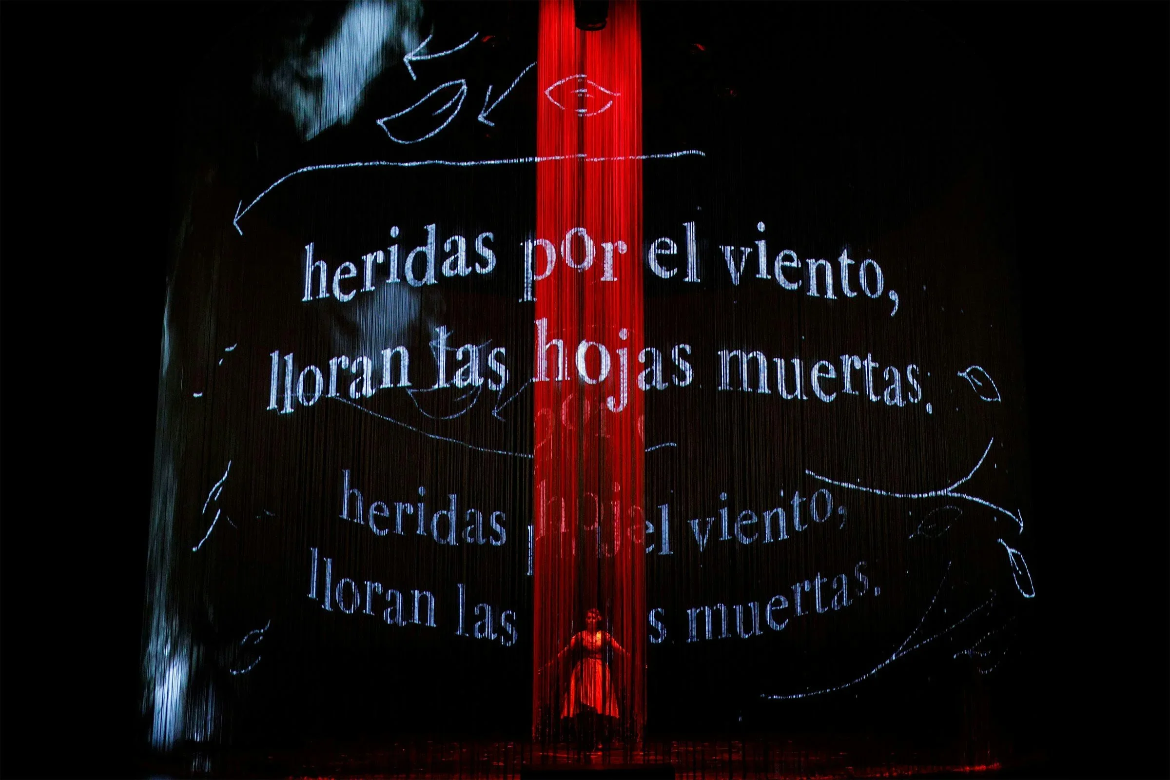 Projection of a quote in Spanish, 'heridas por el viento, lloran las hojas muertas,' with a red vertical stripe down the center and a silhouette of a person at the bottom, surrounded by leafy sketches.